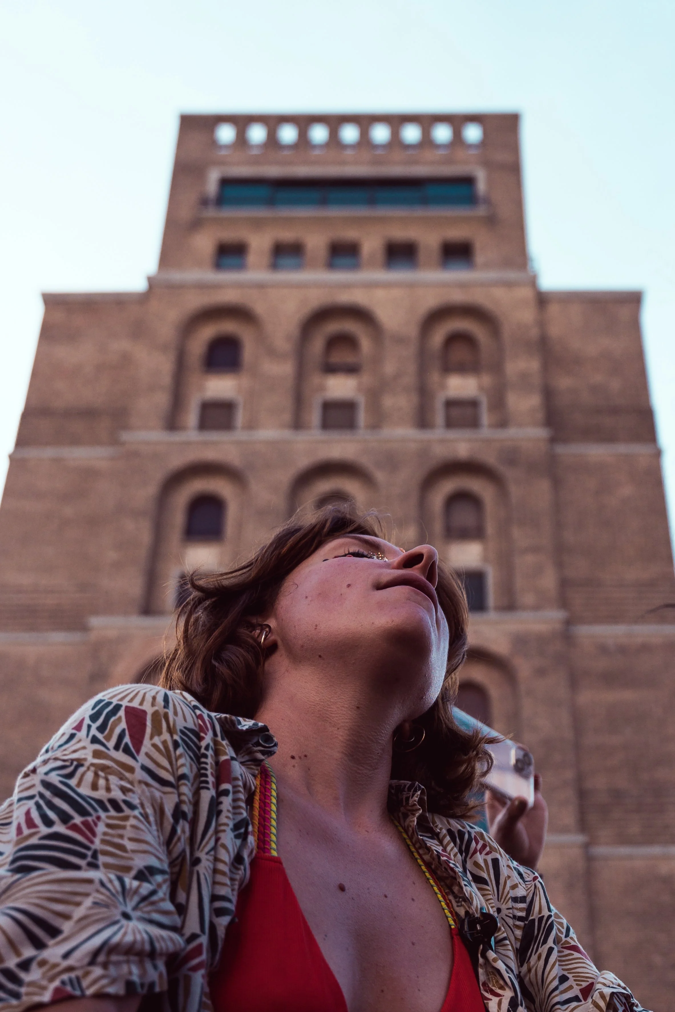 Una donna con i capelli corti e indossa una maglietta rossa, scatta una foto con uno smartphone davanti a un edificio storico in mattoni.