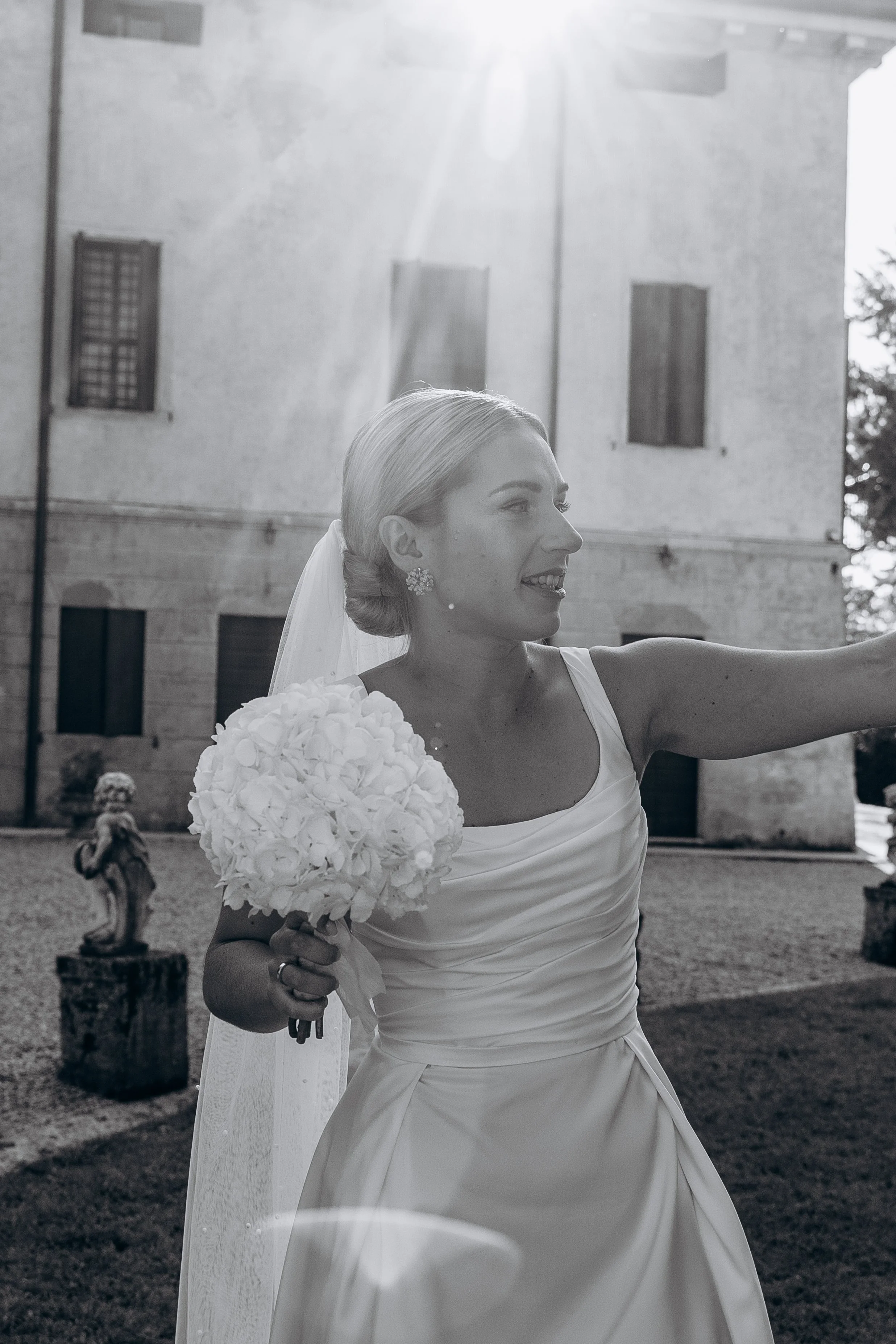 Una sposa sorridente con un bouquet di fiori bianchi in una mano, che si trova all'aperto davanti a un edificio stile storico, con il sole che splende dietro di lei.