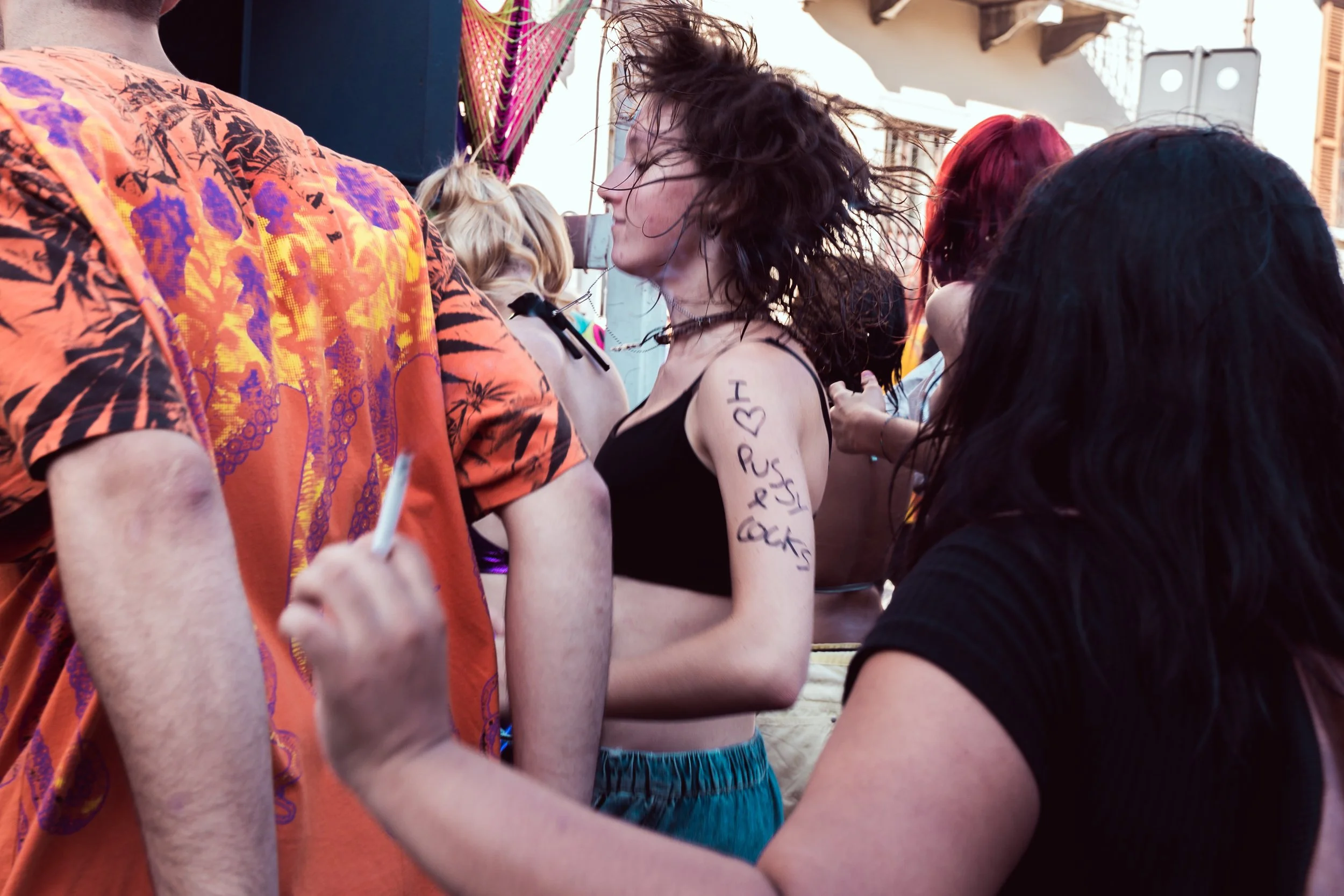 Un gruppo di persone si trova all'aperto durante un evento o una festa, alcune sono mentre si autografi o disegnano. La donna al centro con i capelli mossi e vestita con un top nero e pantaloni corti verdi sorride tra le altre persone, una delle qual