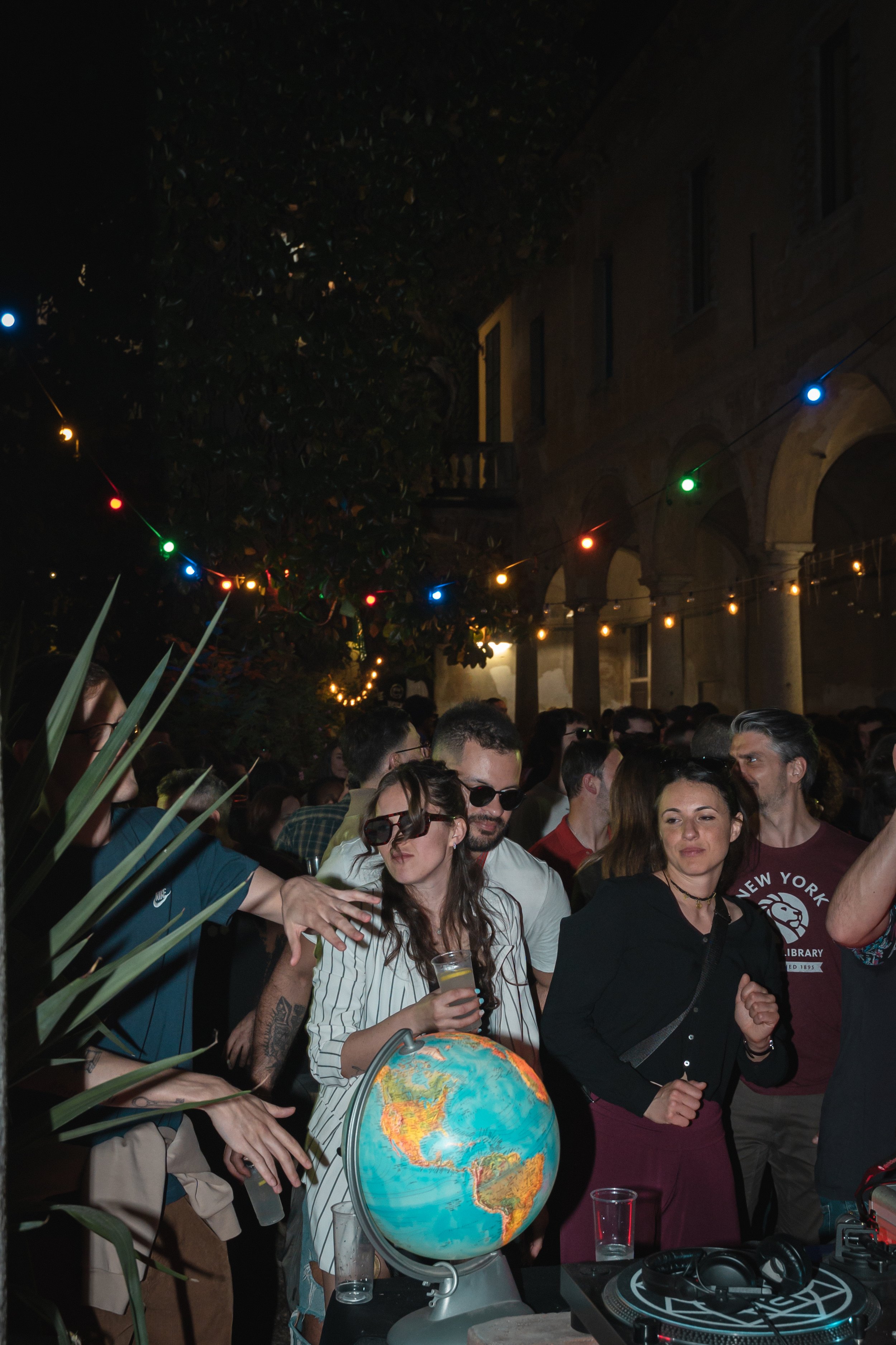 Folla di persone a una festa notturna all'aperto con luci colorate e un globo terrestre su un tavolo.
