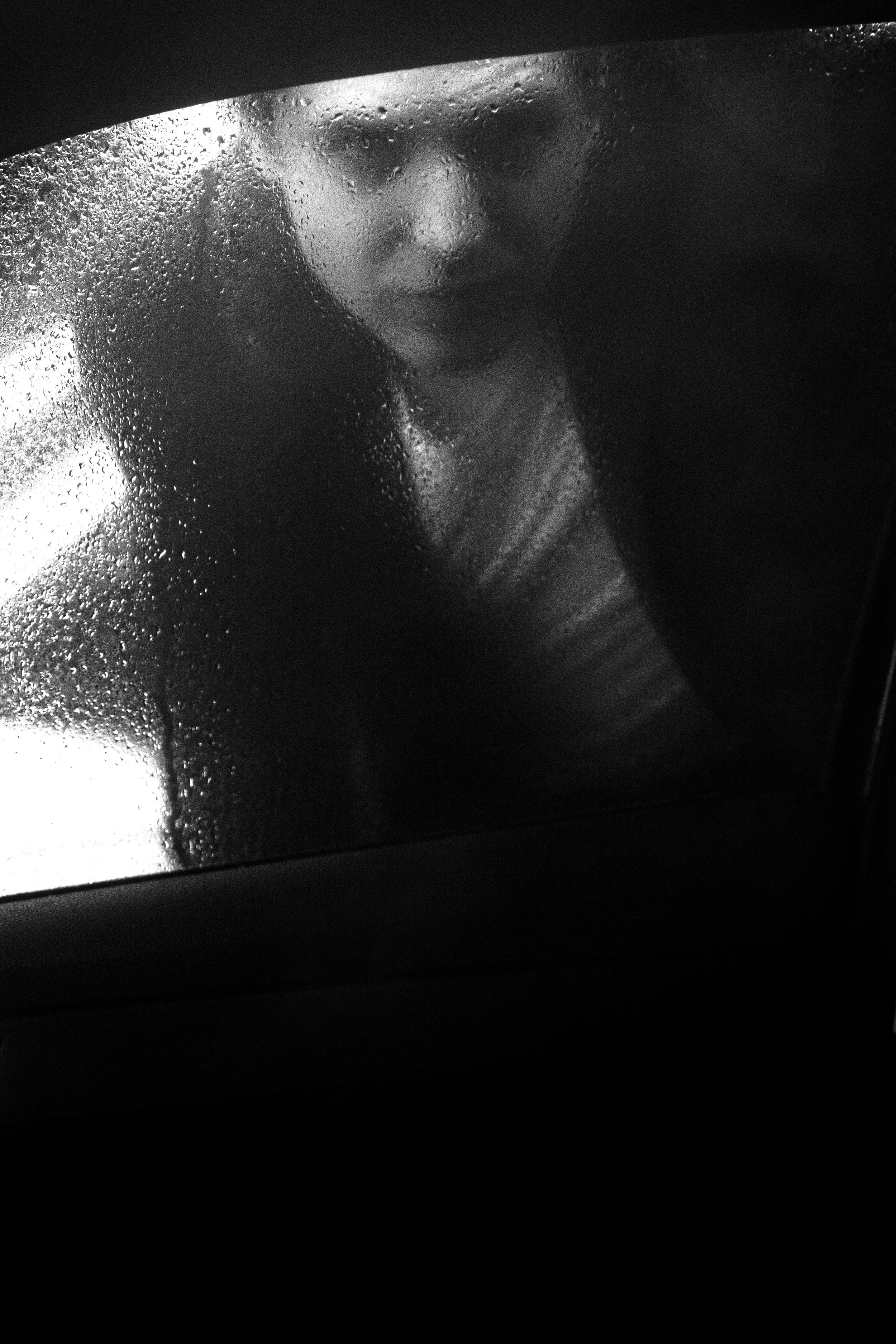 Donna che guarda attraverso una finestra appannata e bagnata, con gocce di pioggia sulla superficie, con un'espressione pensierosa.