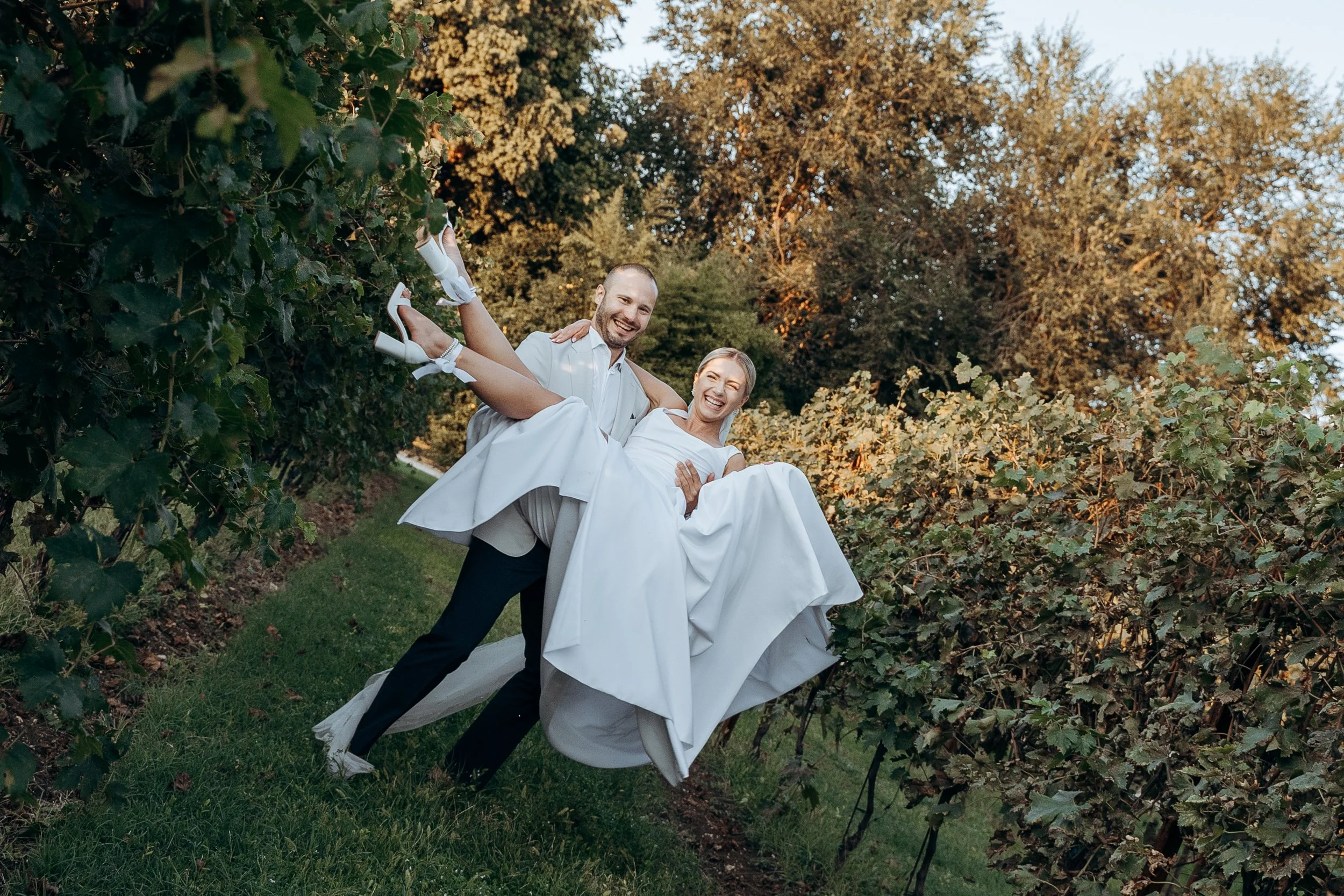 Una coppia di sposi felici, il marito che solleva la sposa, circondati da filari di vite in un vigneto durante il tramonto.