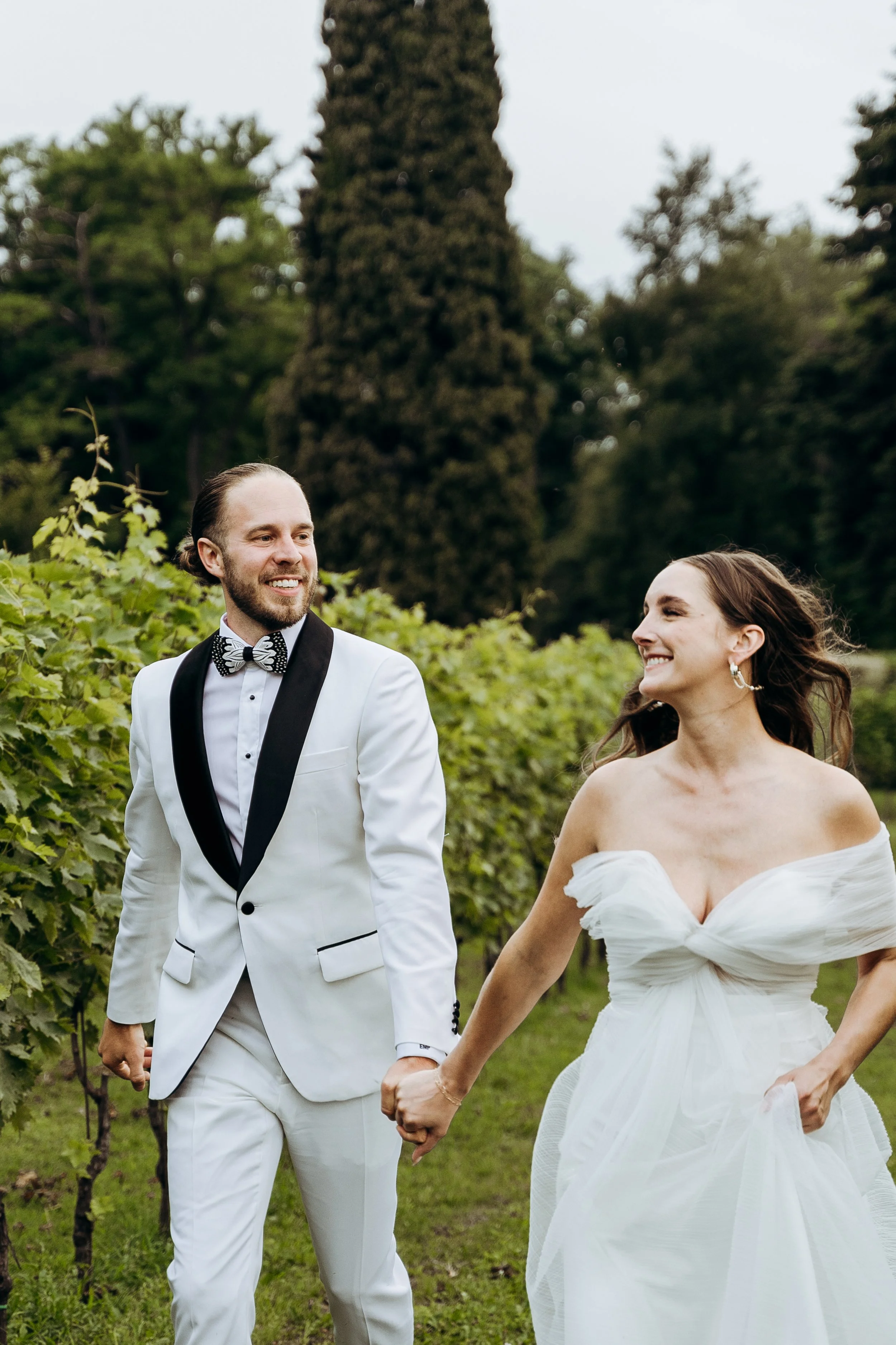 Una coppia di sposi in abiti da matrimonio, felici e sorridenti, che si tengono per mano in un prato verde con alberi sullo sfondo.