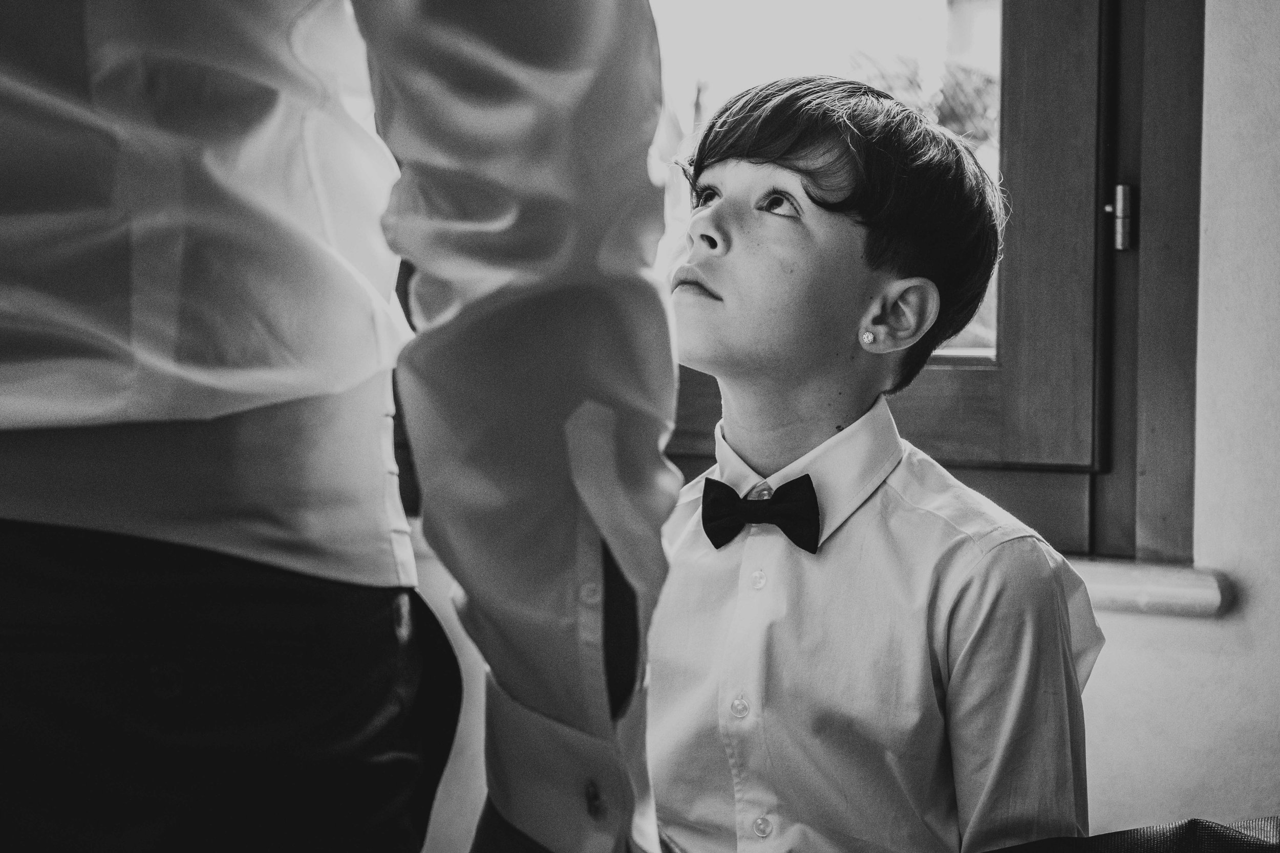 Ragazza con vestito elegante che aiuta un ragazzo con camicia bianca e fiocco nero, seduto vicino a una finestra in un ambiente interno.