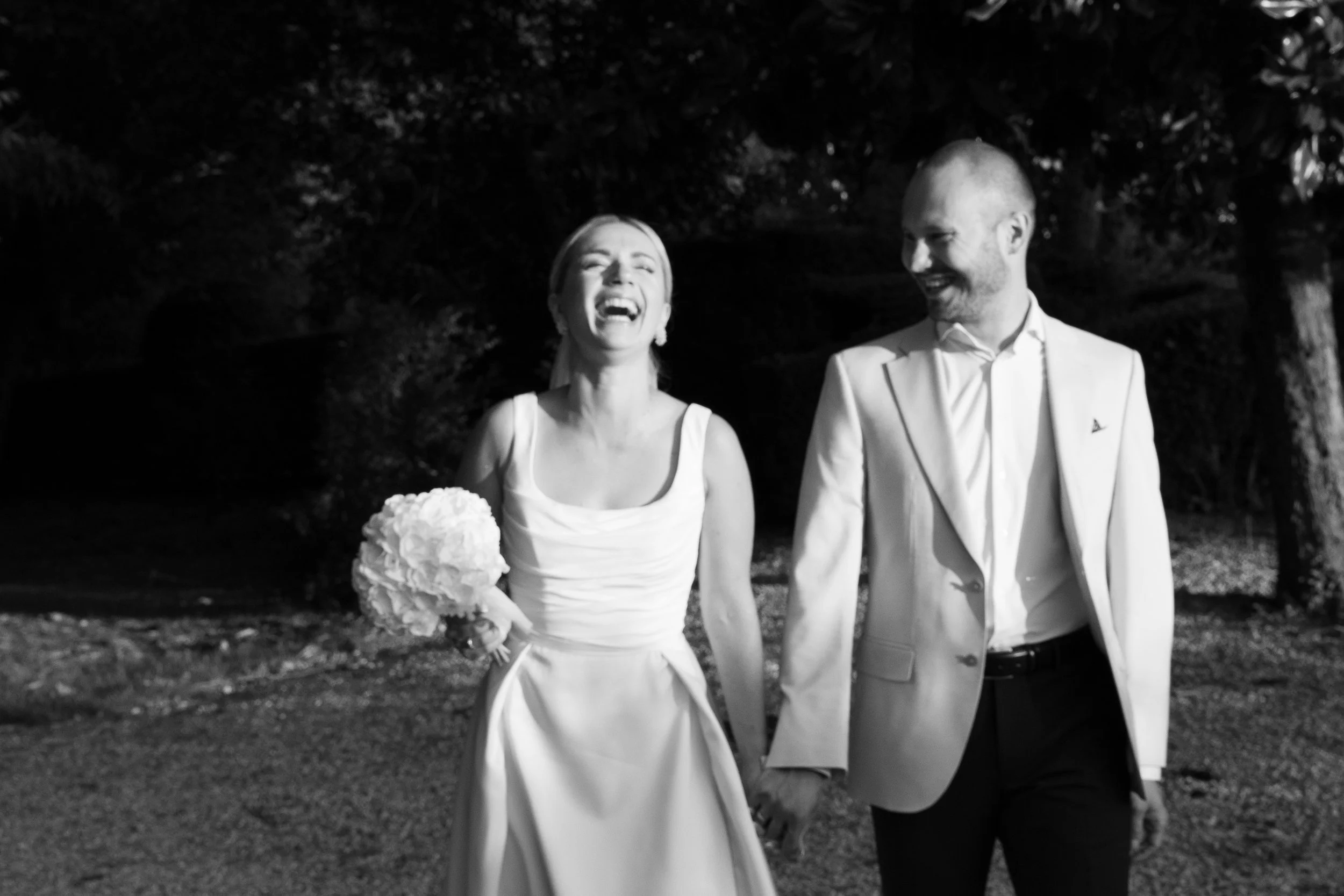 Una coppia di sposi felici cammina mano nella mano all'aperto, lui indossa un abito chiaro e lei un abito da sposa con bouquet, entrambi sorridenti.
