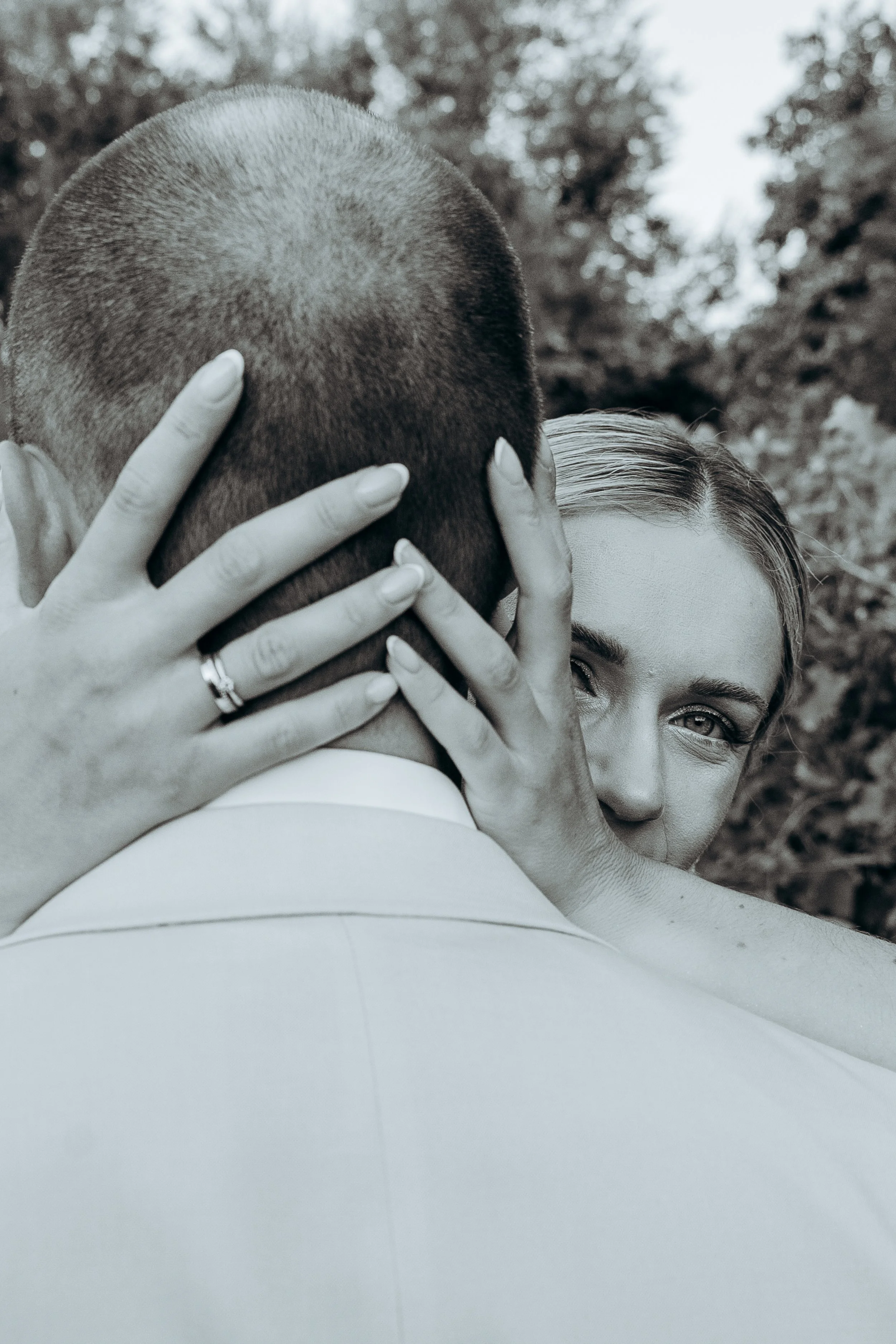 Un uomo e una donna si abbracciano, con il volto della donna che si intravede tra le mani dell'uomo. La donna sorride e guarda verso la fotocamera, in uno sfondo di natura.
