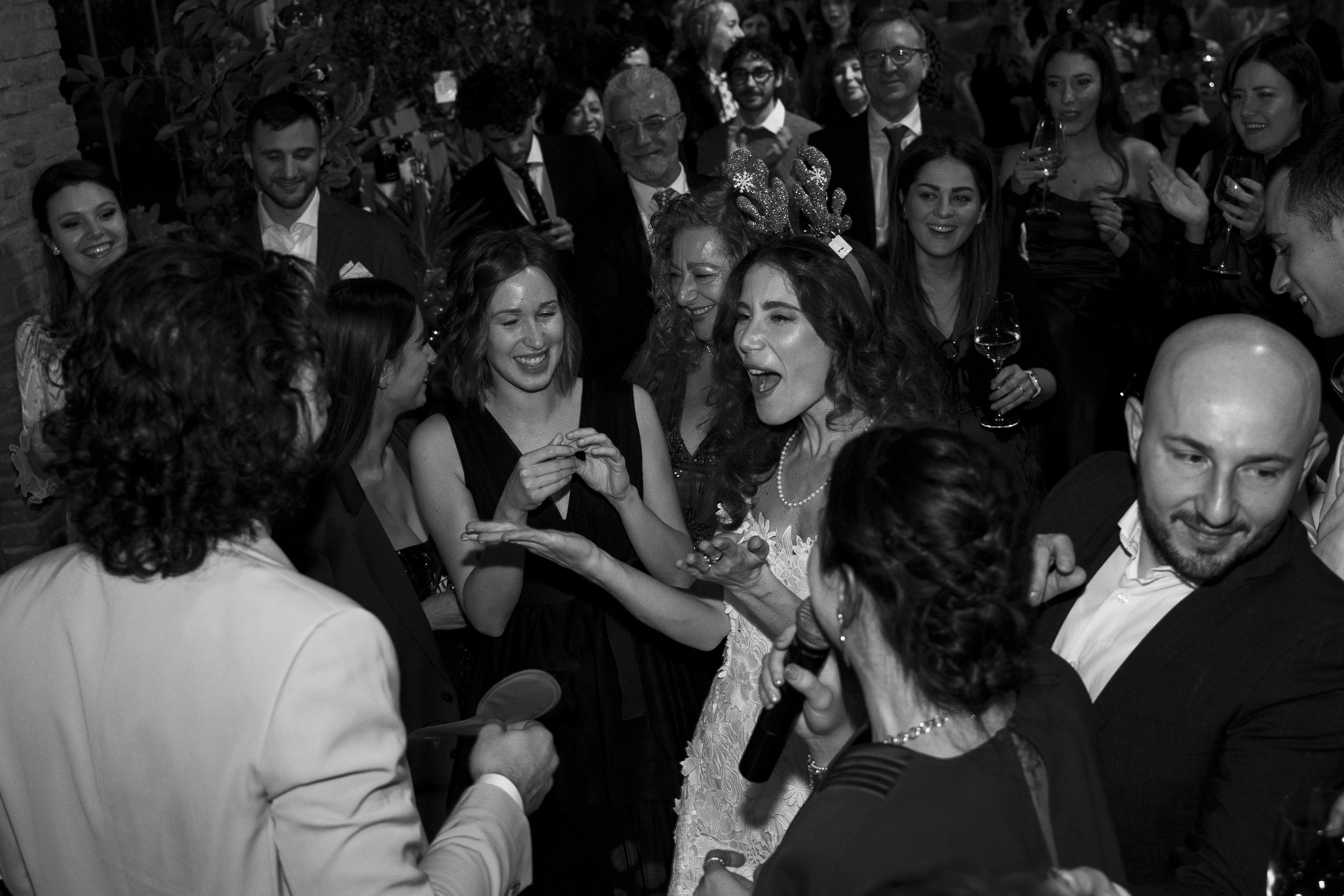 Un gruppo di persone sorridenti e che si divertono durante una festa o un ricevimento, con alcune persone che tengono bicchieri di vino e altre che ridono e chiacchierano, con uno sfondo di luci e decorazioni festive.