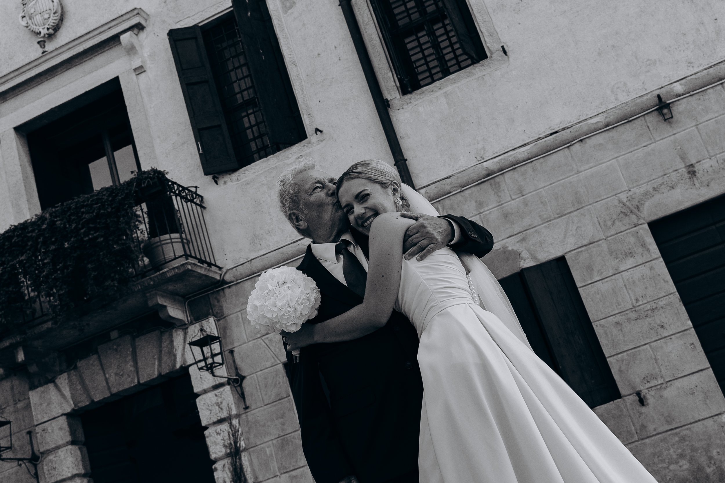 Un uomo anziano bacia una donna in abito da sposa sulla guancia, sullo sfondo di un edificio. La donna sorride felice e tiene un bouquet bianco.