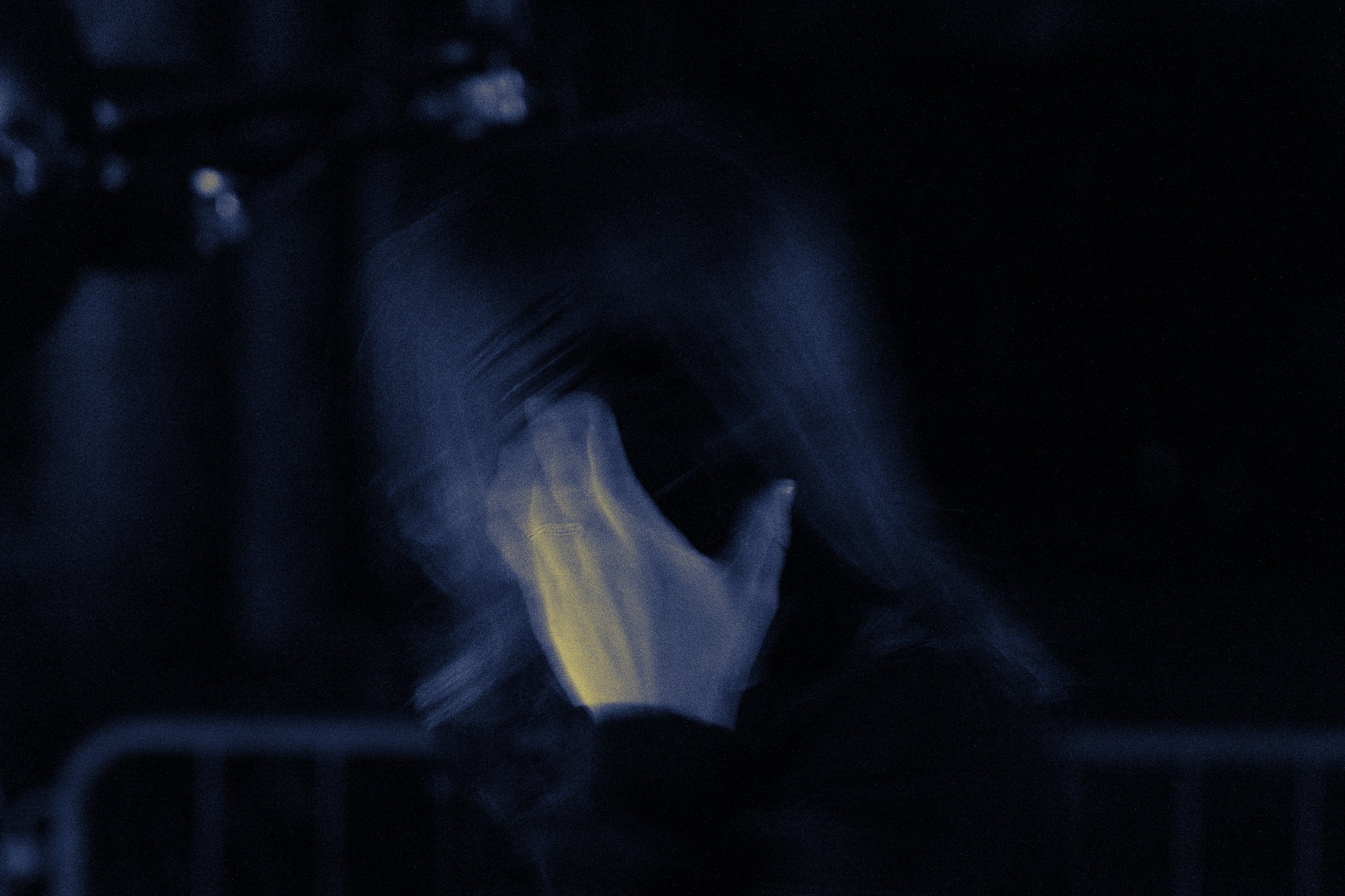 Una persona con lunghi capelli scuri si copre il volto con una mano, in un ambiente molto oscuro, con poca luce che crea un effetto misterioso e sfocato.