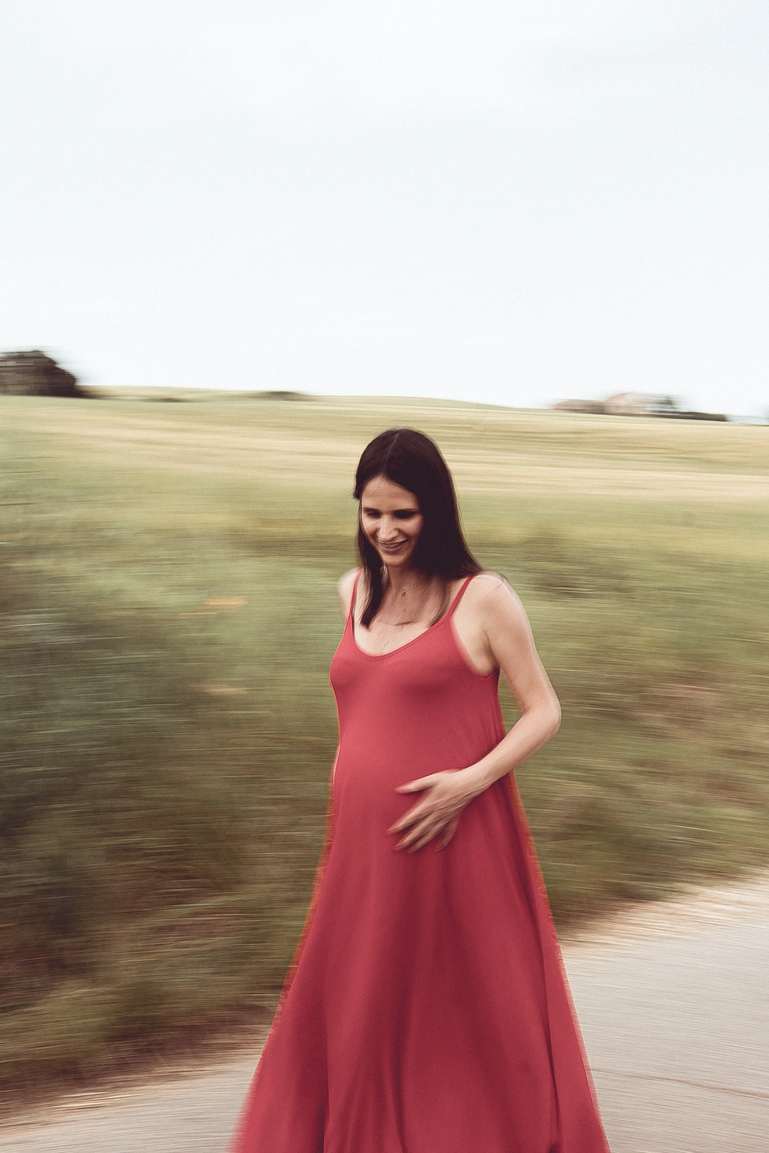Una donna in vestito lungo rosso cammina all'aperto su un sentiero con campagna verde sullo sfondo, sorridendo.