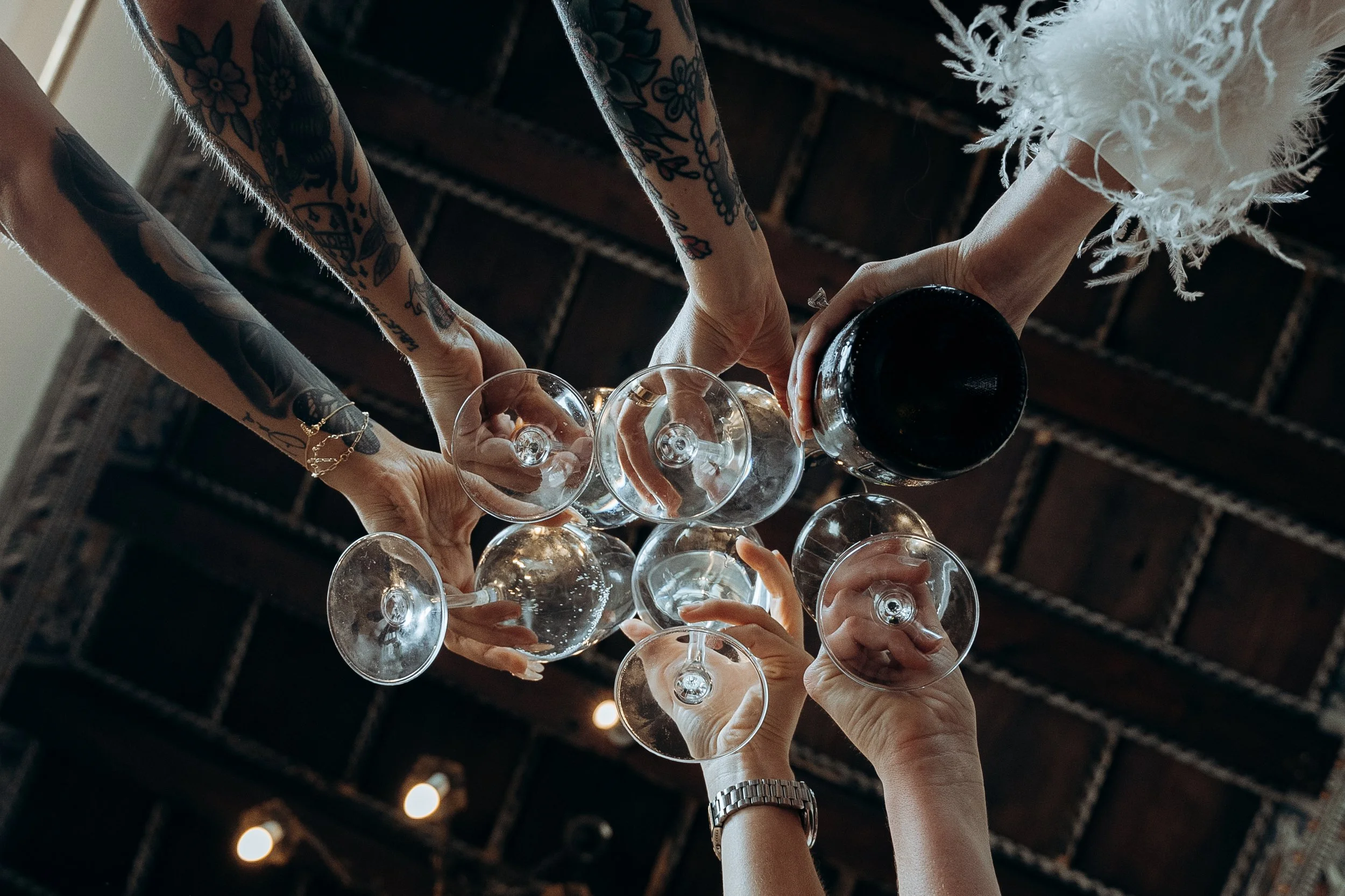 Persone che brindano con bicchieri di vino, alcuni con tatuaggi e uno con orologio, in un ambiente con soffitto in legno.