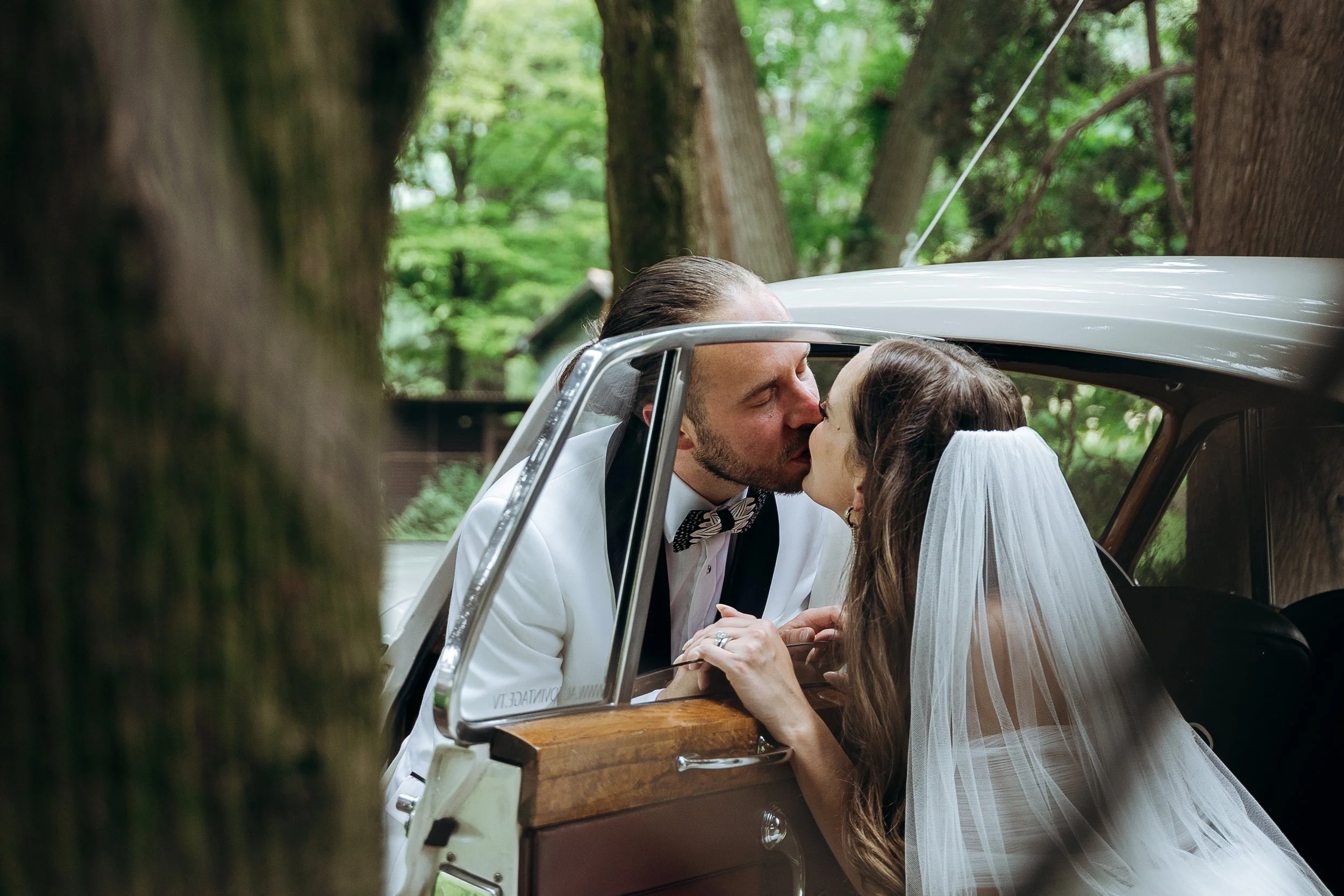 Una coppia di sposi si bacia in auto mentre si trova in un bosco, con alberi verdi sullo sfondo.