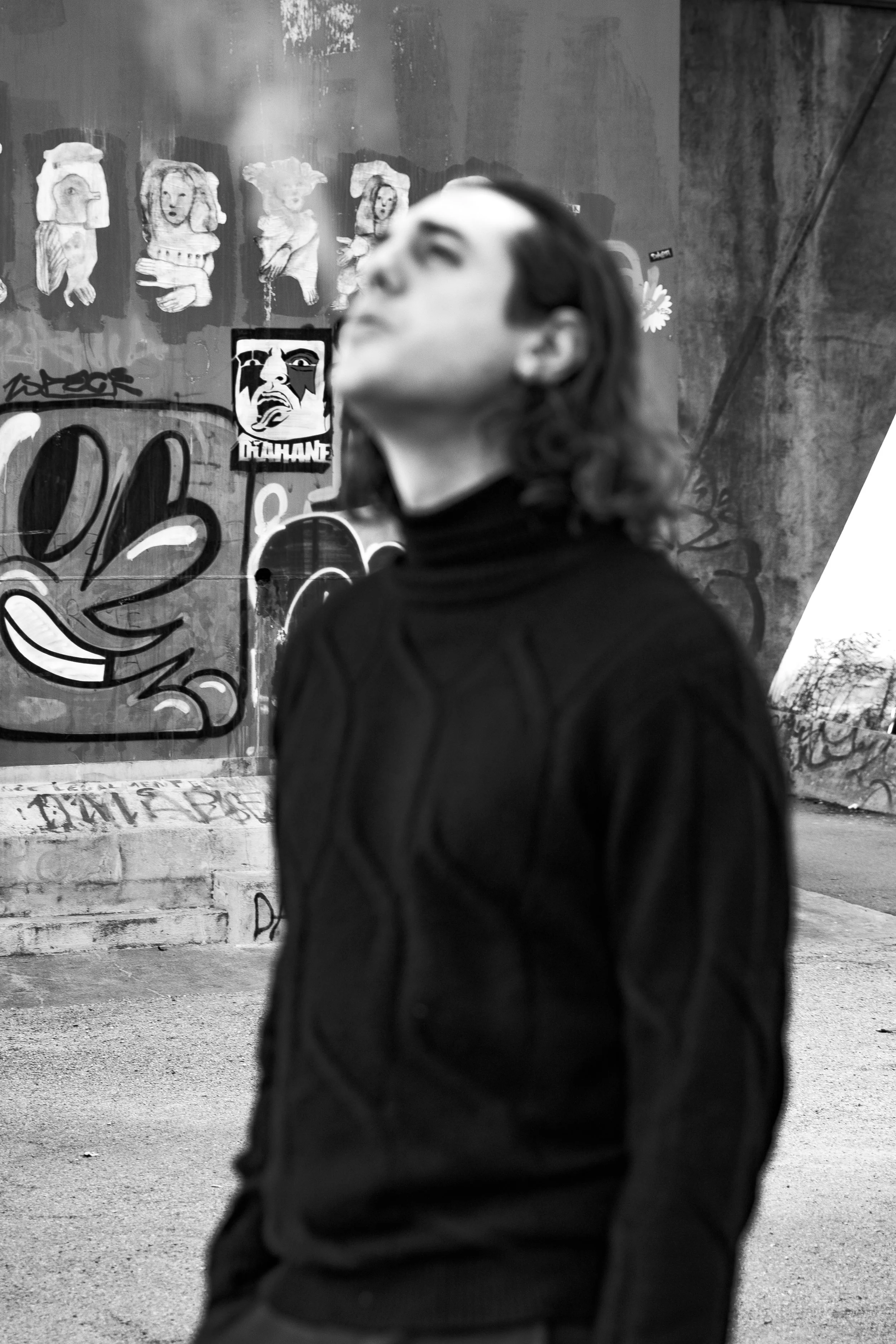 Persona con capelli lunghi e vestito nero in primo piano, sfondo con graffiti e poster su un muro urbano.