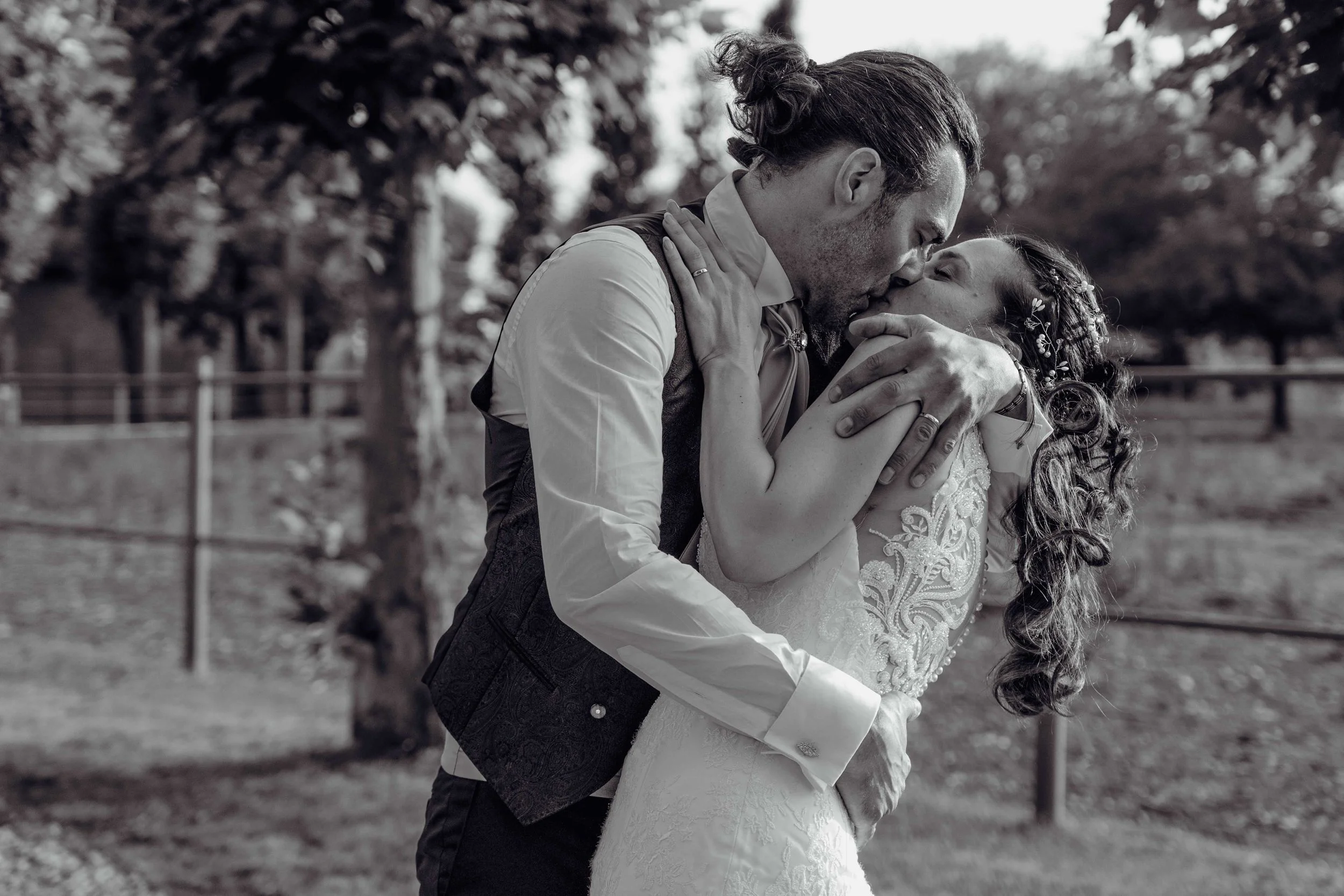 Una coppia si bacia appassionatamente all'aperto, probabilmente durante un matrimonio, con alberi sullo sfondo.