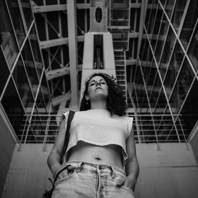 Una donna con capelli ricci scuri indossa una maglietta corta e jeans, in piedi sotto una struttura architettonica moderna di vetro e acciaio, vista dall'alto.