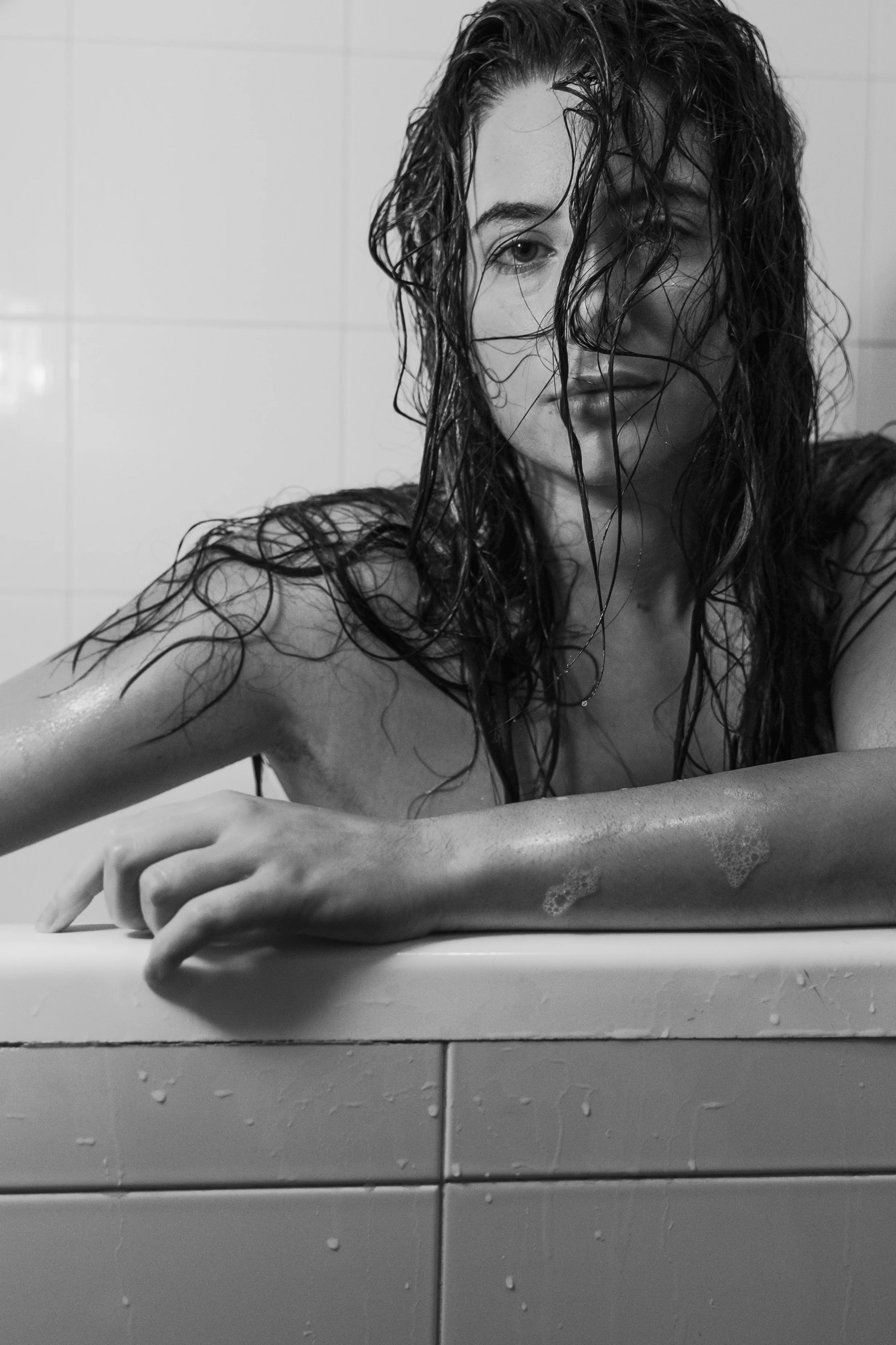 Donna con capelli bagnati, poggiata su una vasca da bagno di piastrelle, con parte del volto coperta dai capelli e il braccio appoggiato sulla vasca.