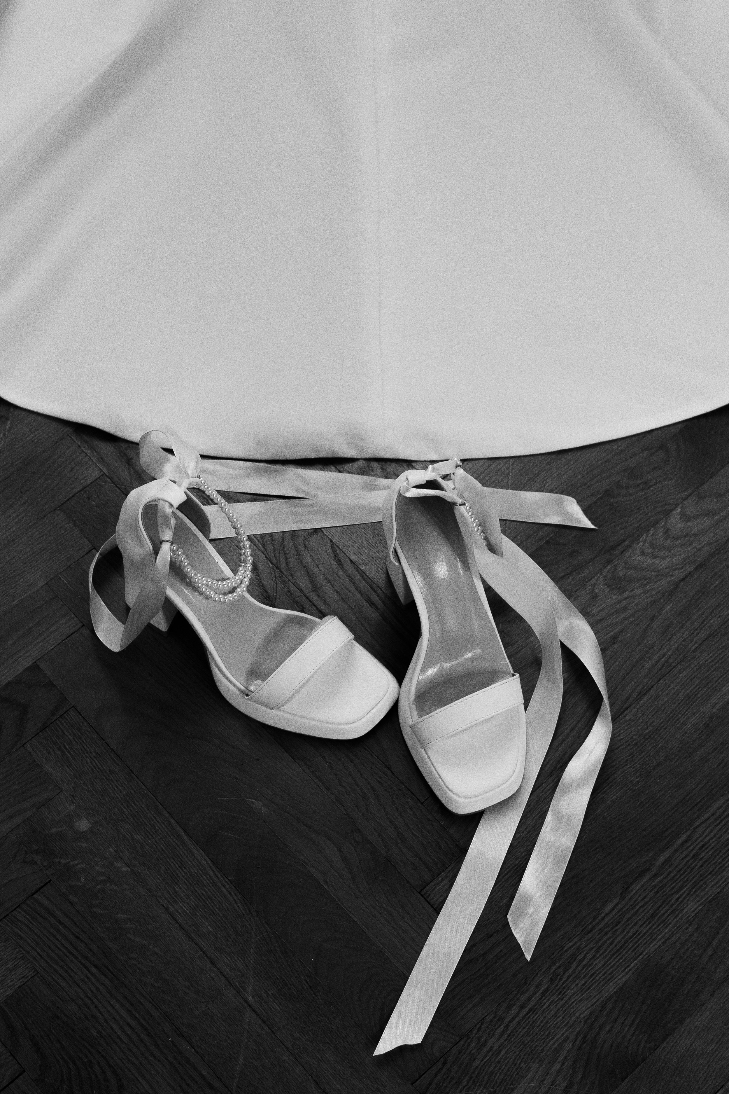 Scarpe da sposa con tacco alto, decorazioni di nastri e perle sul pavimento di legno vicino a un vestito da nozze