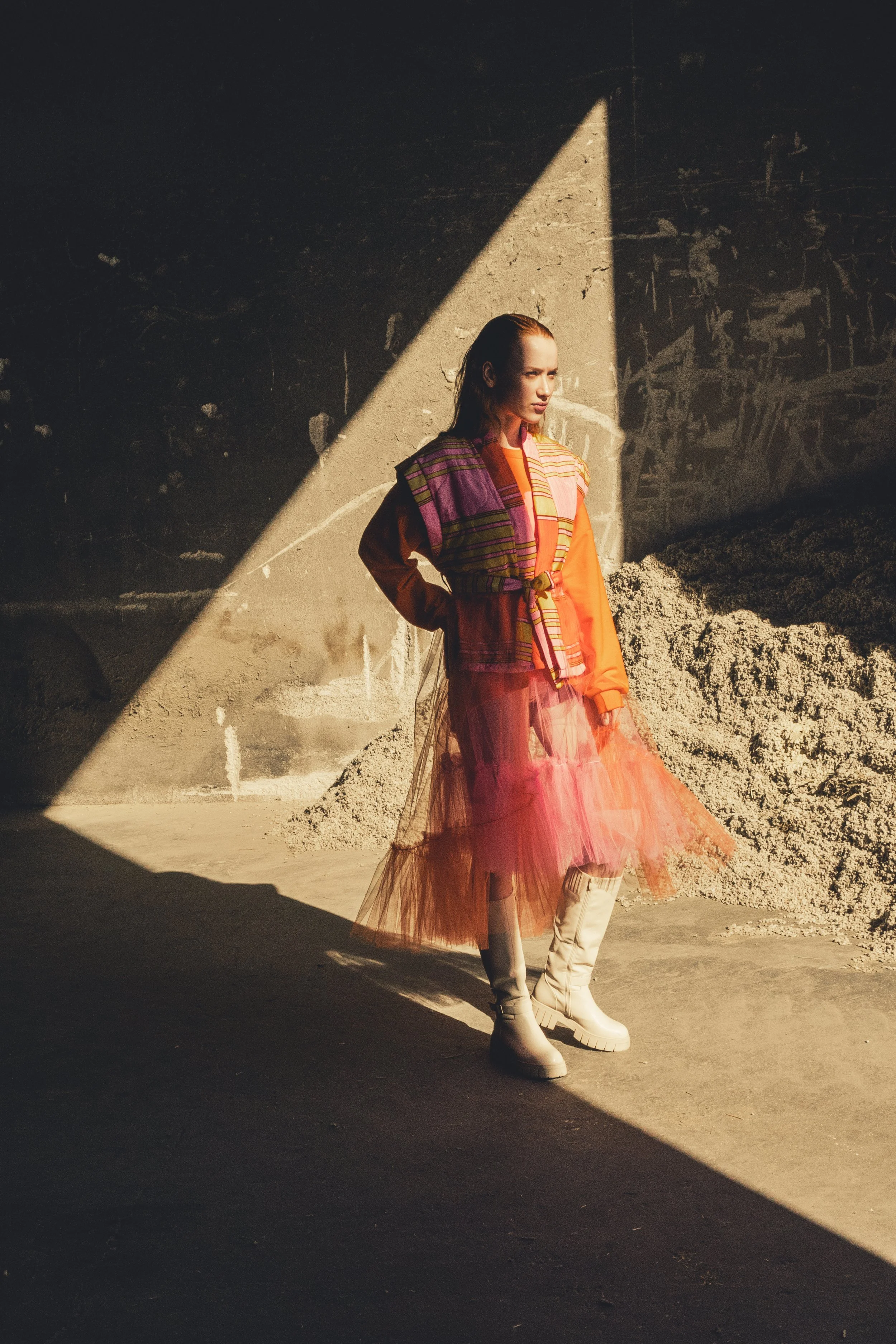 Una donna vestita con abiti colorati e stivaletti bianchi si trova in un ambiente con pareti di cemento e una pila di sabbia, illuminata da una luce che crea un contrasto con l'ombra.