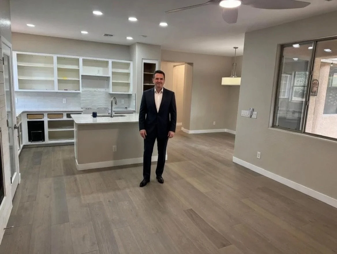 A man in a suit standing inside an empty, modern kitchen and living area with hardwood floors, white cabinets, and large windows.