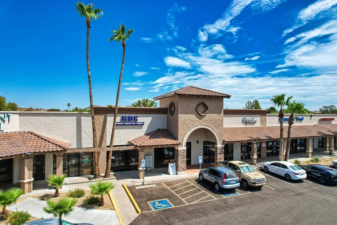 A strip mall with parking lot, a few palm trees, and storefronts including R.D.C., Queens of Kings, and Tom Thumb under a blue sky with scattered clouds.
