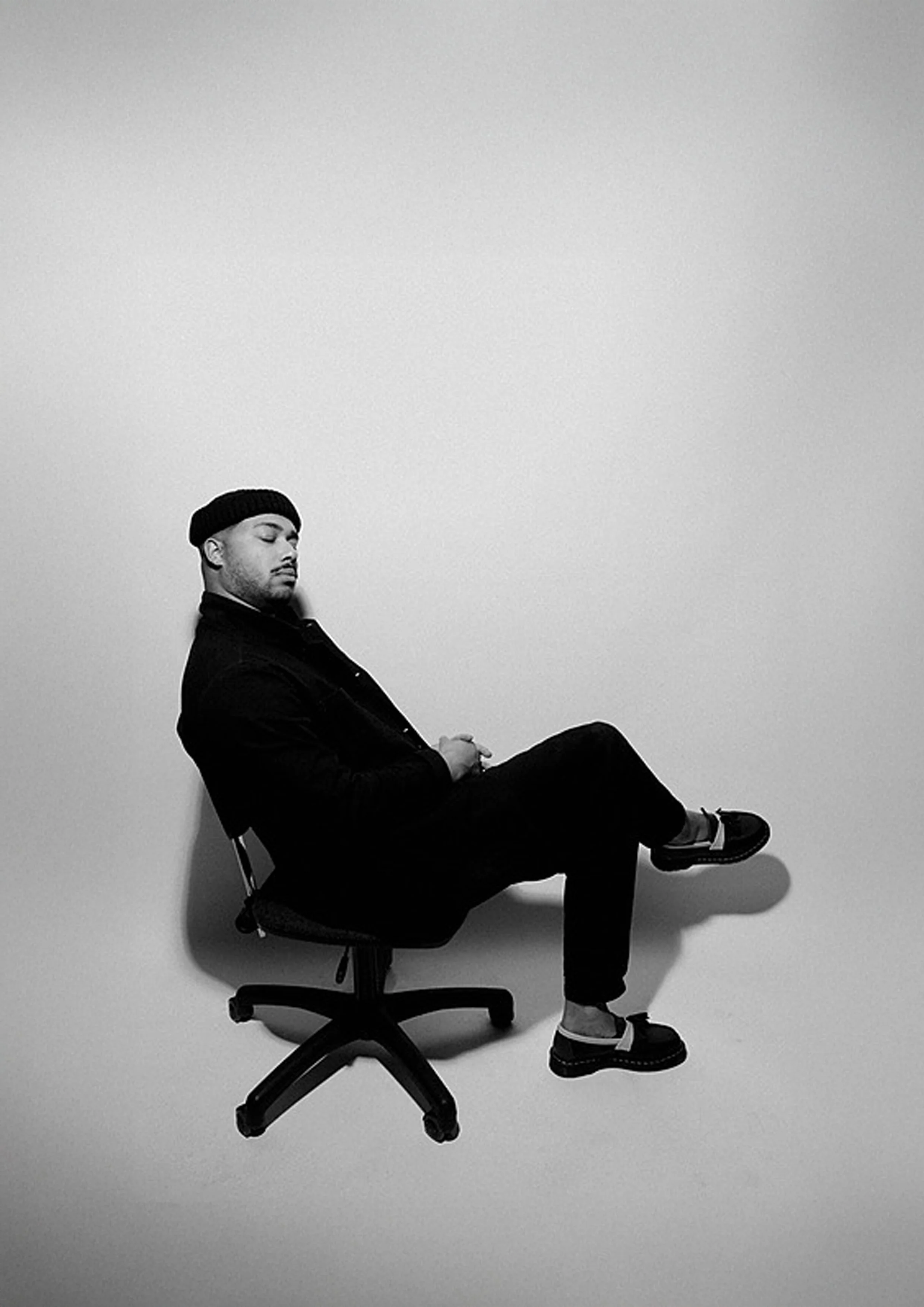 A black and white photo of a young man sitting on a rolling office chair, with his eyes closed, wearing a beanie, dark jacket, dark pants, and sneakers, against a plain light background.