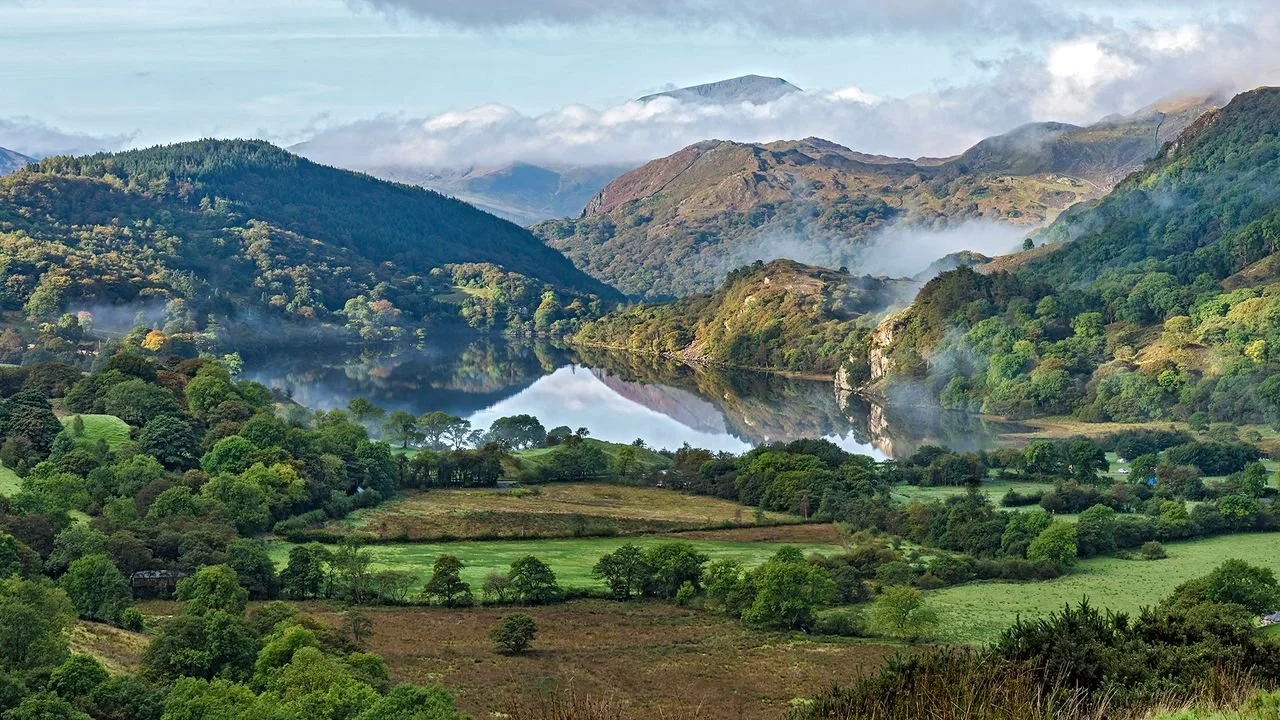 Scenic view of a mountain landscape with a lake reflecting the mountains and sky, surrounded by lush green trees and grass.
