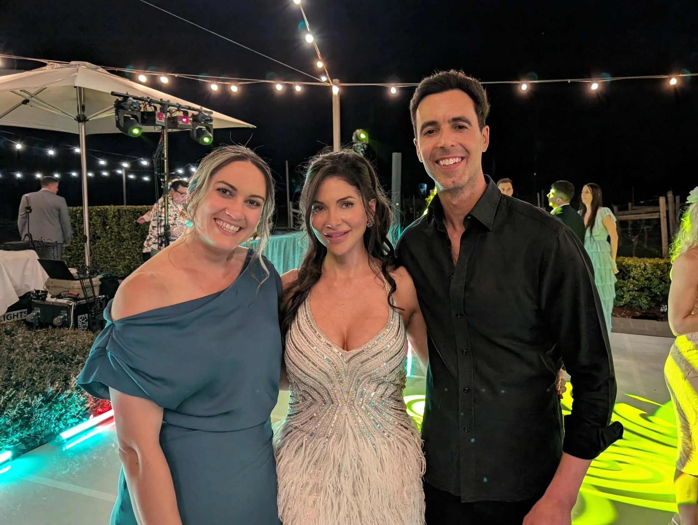 Three people smiling at a night outdoor event, with string lights and a DJ setup in the background.