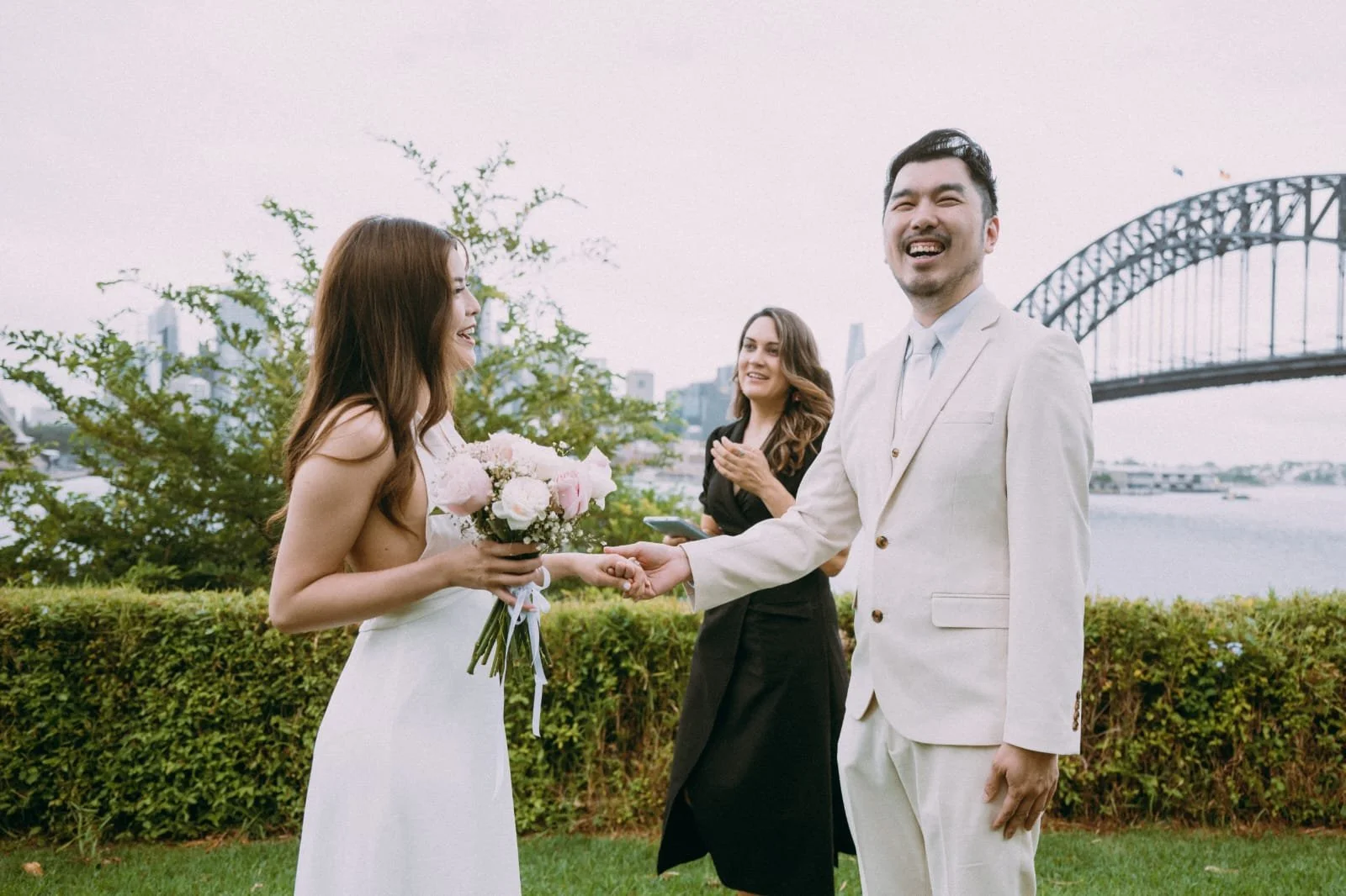 A couple exchanging wedding vows outside near the water, with a ferris wheel in the background. The bride is holding a bouquet of flowers, and the groom is smiling in a white suit. A woman officiant stands behind them, clapping.