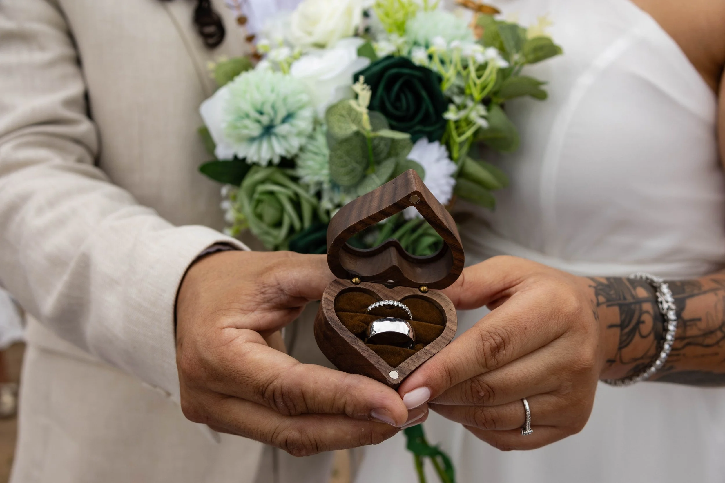 Person holding a heart-shaped wooden jewelry box with a wedding ring and diamond ring inside, standing in front of a bouquet of white and green flowers.