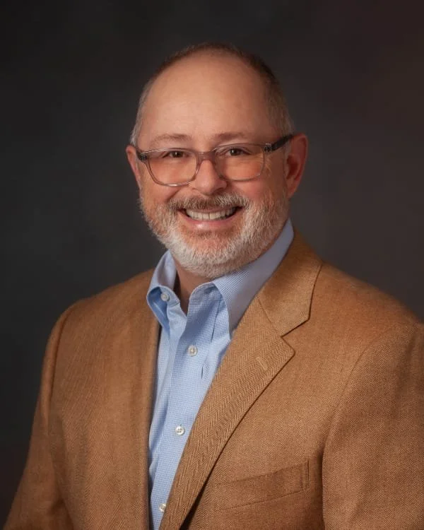 A smiling middle-aged man with glasses, a beard, and short hair dressed in a light blue shirt and a tan blazer, posing against a dark background.