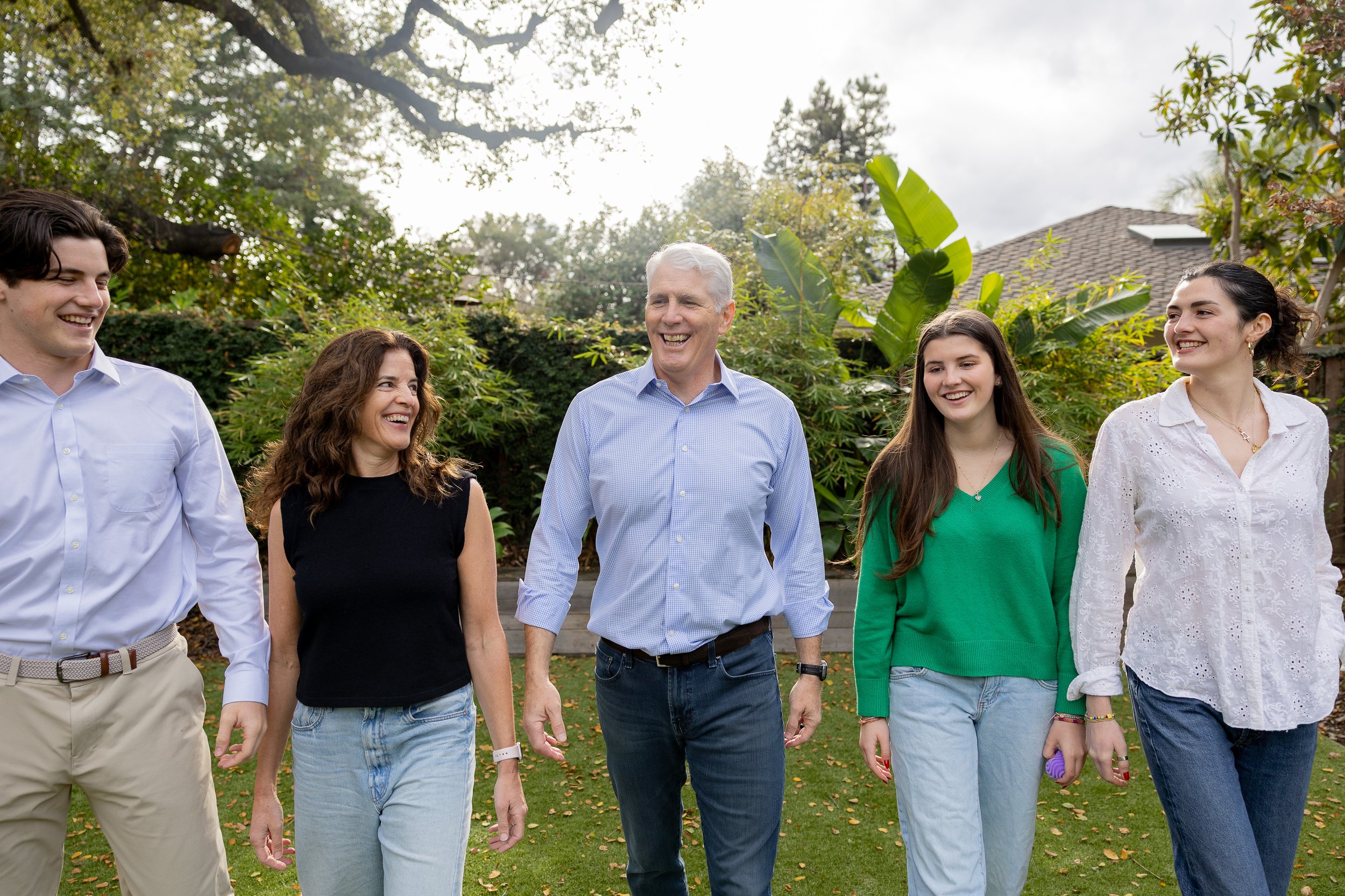 People smiling and walking outdoors in a garden or backyard setting with greenery and trees.