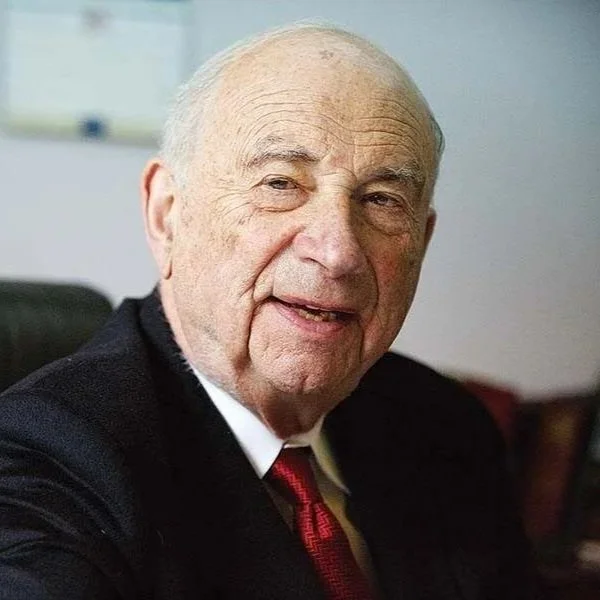 Older man with a mild smile, wearing a black suit and red tie, sitting in an office environment.