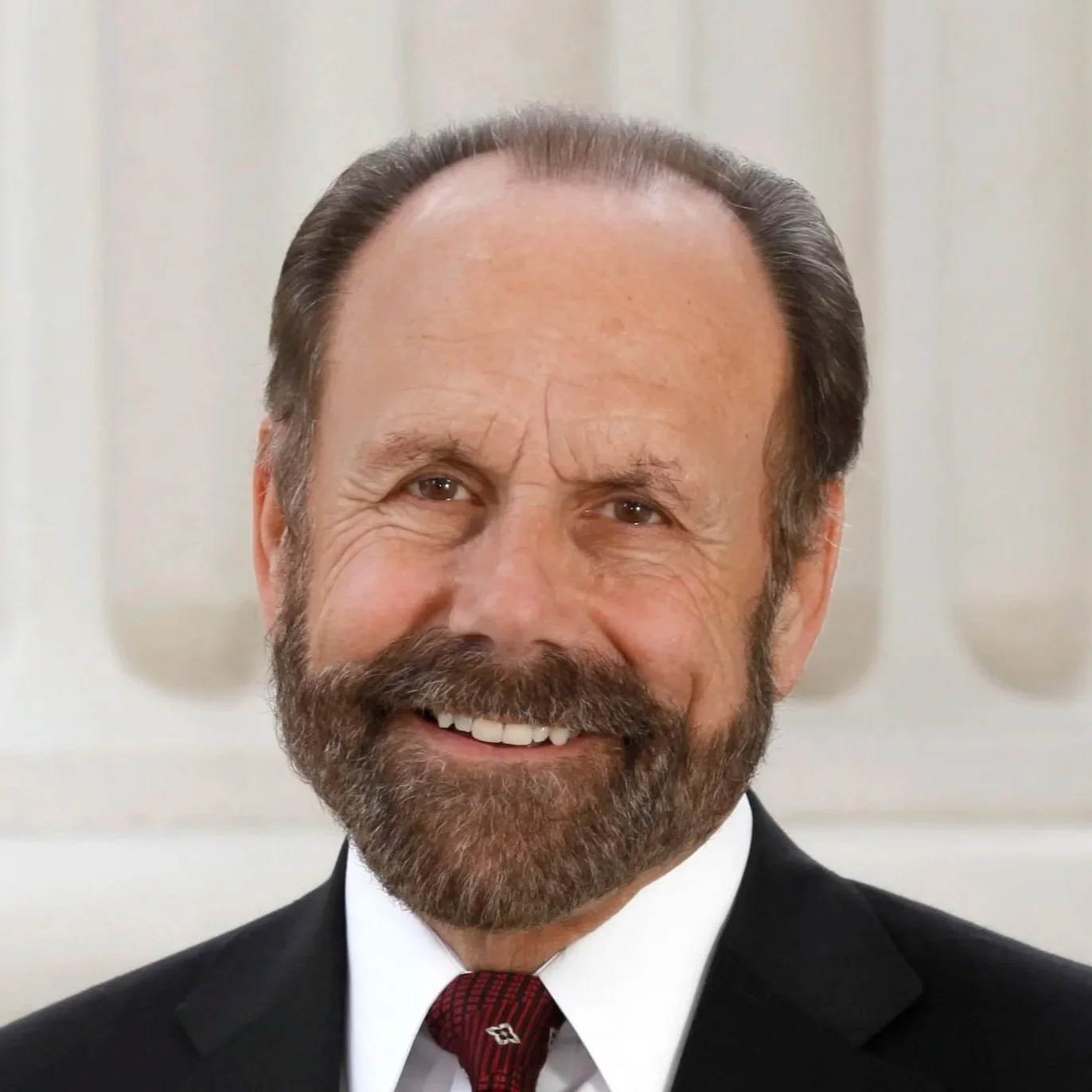 A middle-aged man with a beard and mustache, dressed in a black suit, white shirt, and red tie, smiling, with a background of white molding or architecture.