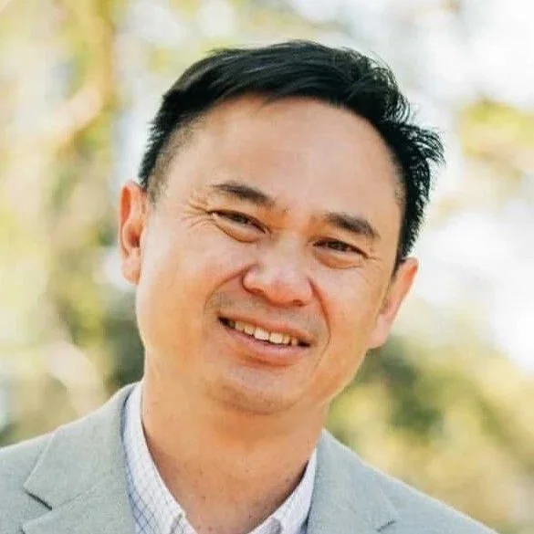 A man with black hair, smiling, wearing a light gray blazer and a checkered shirt outdoors with blurred trees in the background.