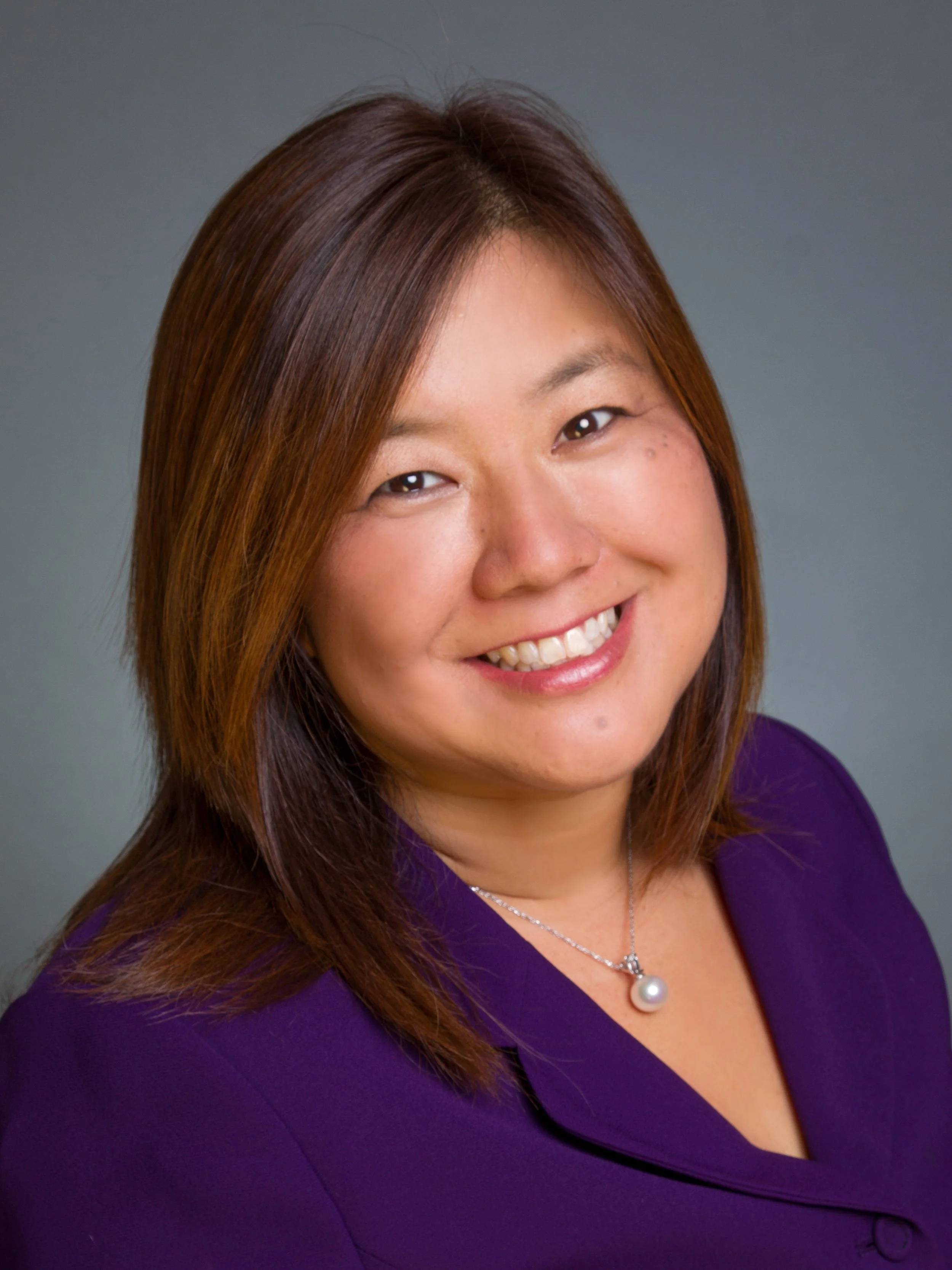 Portrait of a smiling woman with shoulder-length brown hair, wearing a purple blazer and a pearl necklace against a gray background.