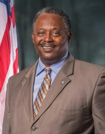 A smiling man in a brown suit, light blue shirt, and plaid tie, standing in front of an American flag.