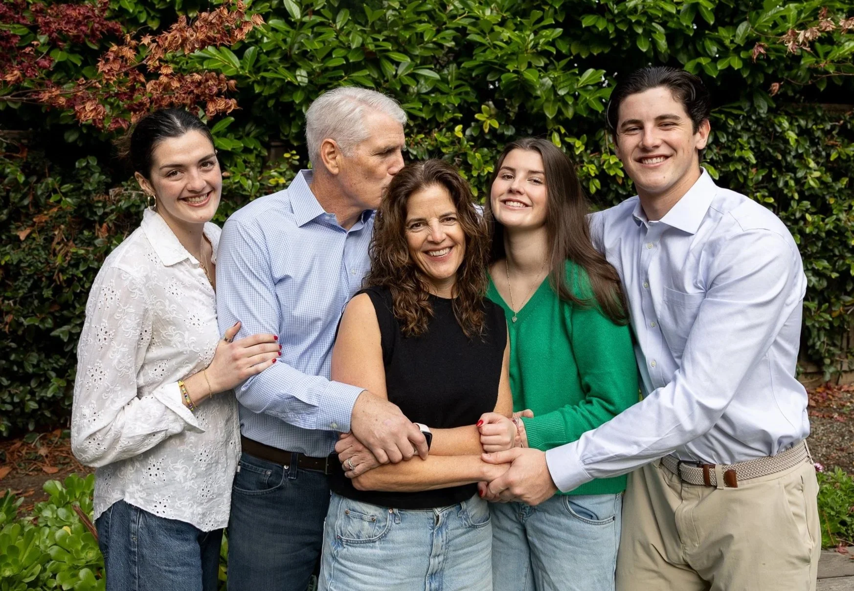 A multi-generational family group standing outdoors, hugging, smiling, and posing for a photo in front of lush green foliage.