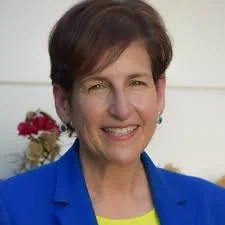 Portrait of a smiling woman with short dark hair, wearing a blue blazer and a yellow top, standing outdoors with flowers in the background.