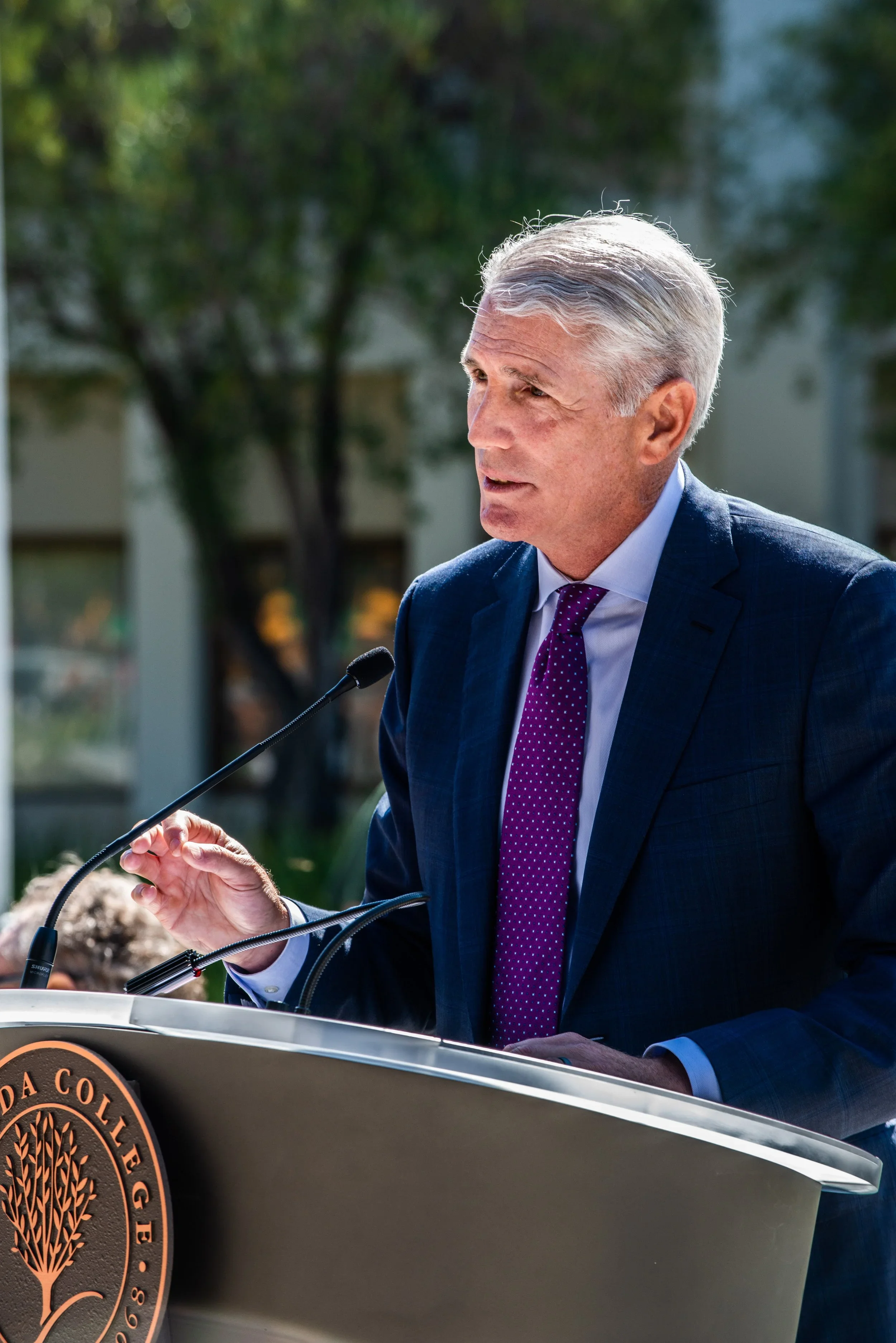 A man in a business suit speaking at an outdoor podium with a university seal, with trees and a building in the background.
