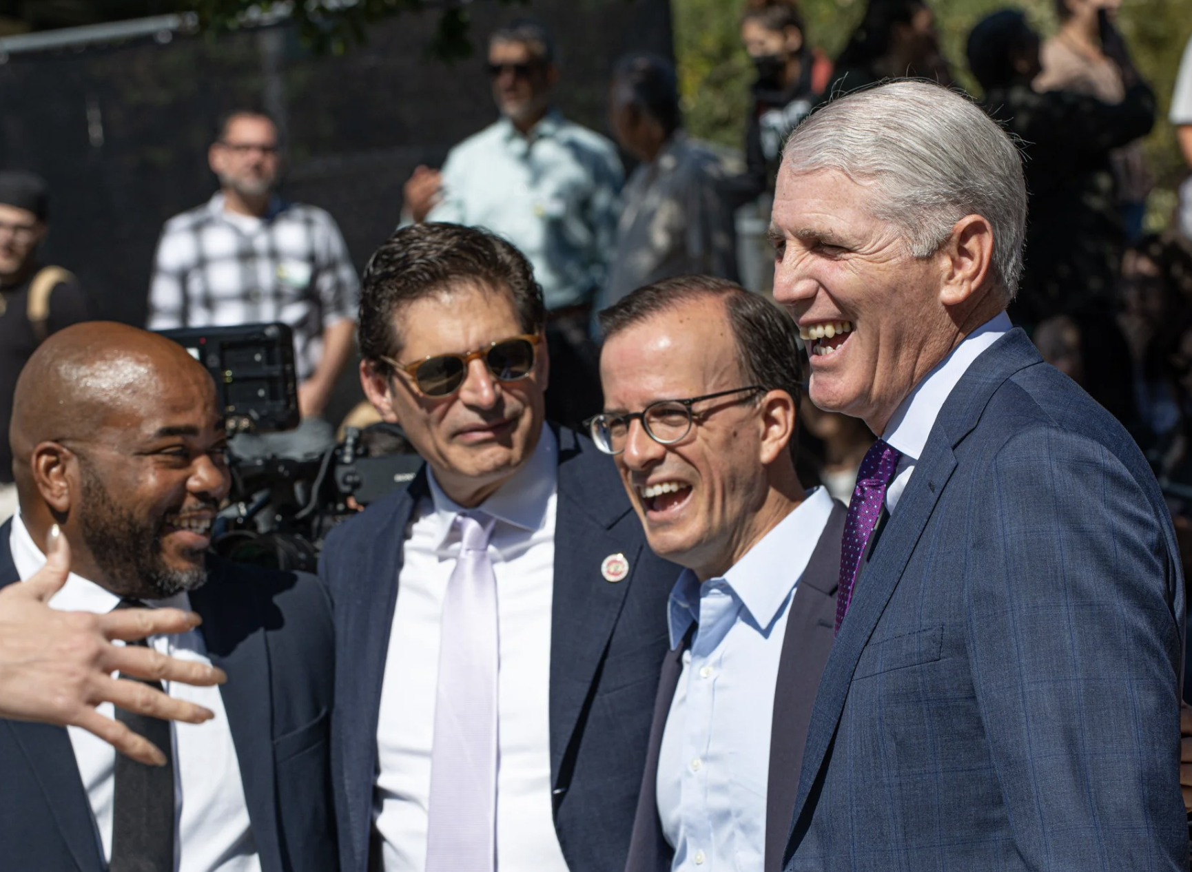 A group of five men in suits and ties laughing and talking together outdoors, with a crowd of onlookers in the background.