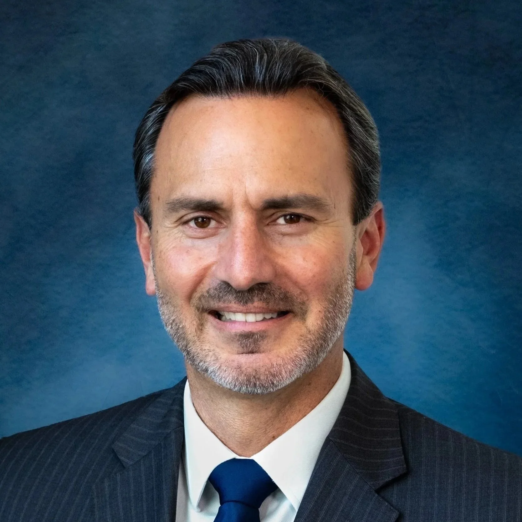 A middle-aged man with dark hair, gray beard, dressed in a dark pinstripe suit, white shirt, and blue tie, smiling against a blue background.