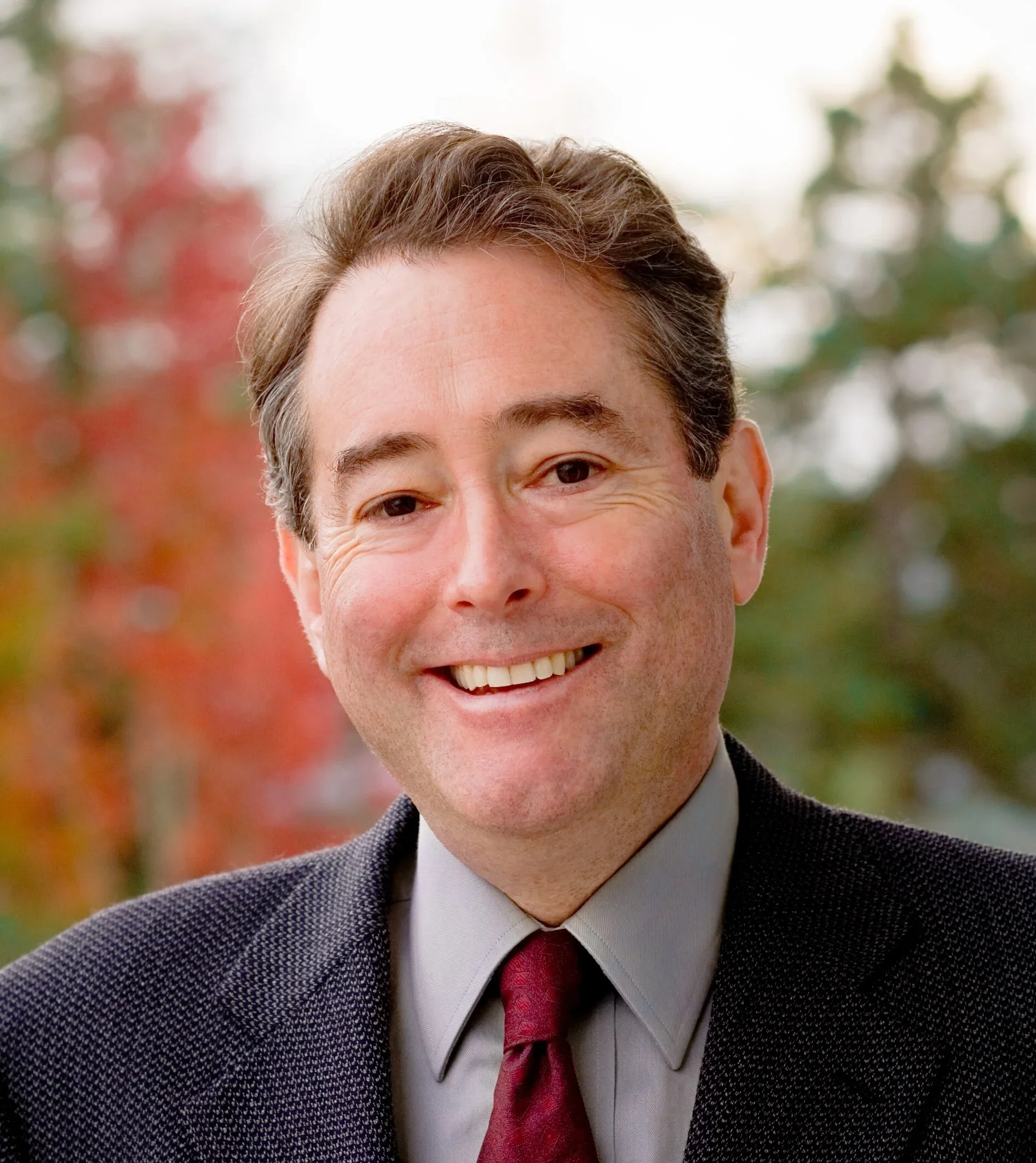 Photograph of a smiling man with brown hair, wearing a dark suit, light gray shirt, and red tie, outdoors with autumn-colored trees in the background.