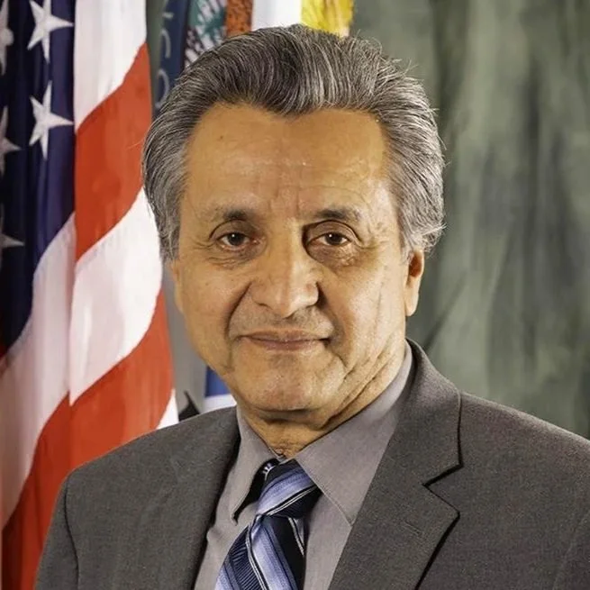 A middle-aged man in a gray suit and tie standing in front of American flags.