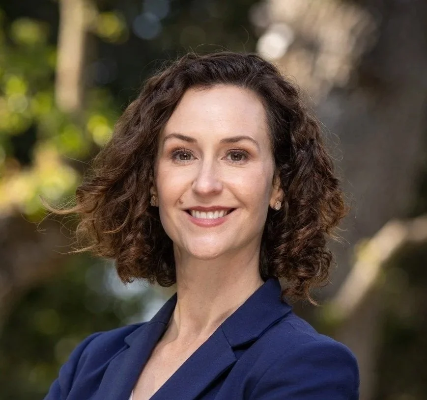 A woman with curly brown hair, wearing a navy blazer, smiling outdoors with blurred trees in the background.