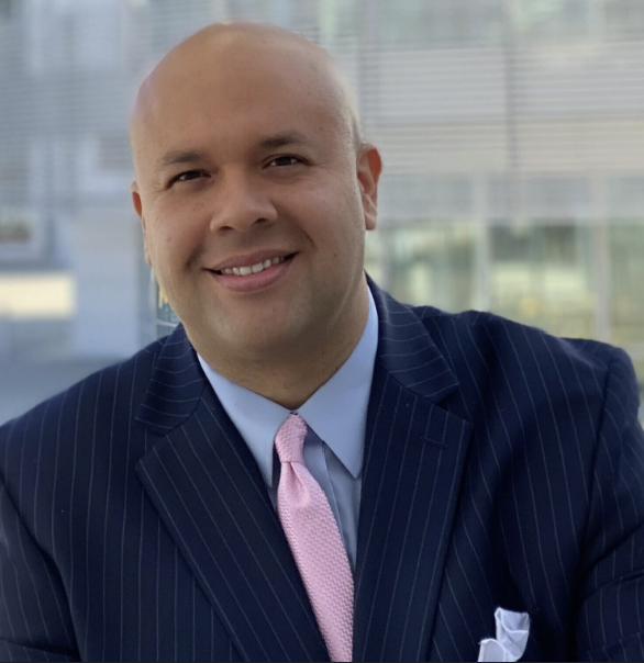 A man with a bald head smiling, dressed in a dark pinstripe suit, light blue shirt, and pink tie, sitting in an office with large windows showing a cityscape background.