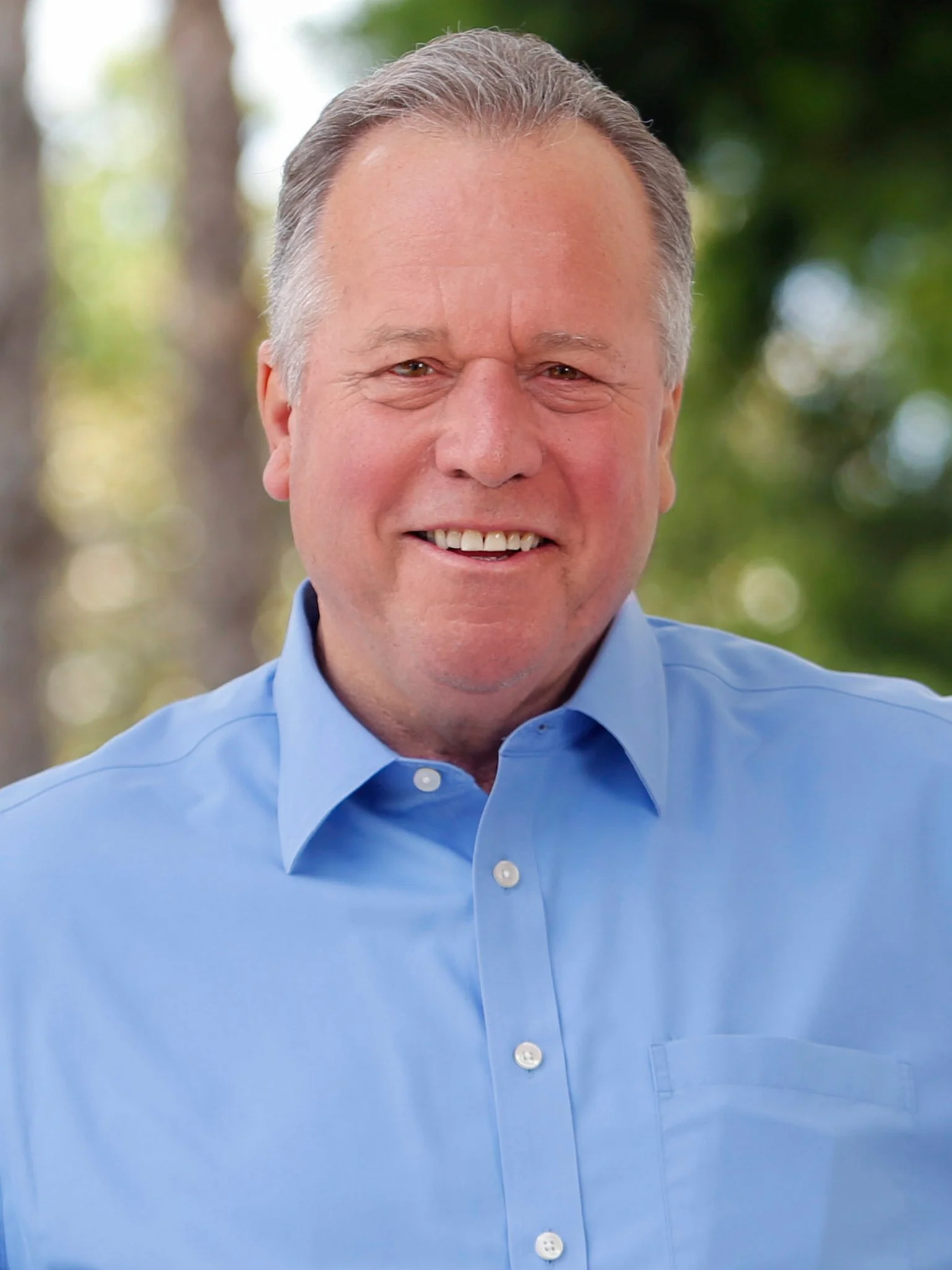 Smiling man with gray hair wearing a blue collared shirt outdoors with blurred trees in the background.