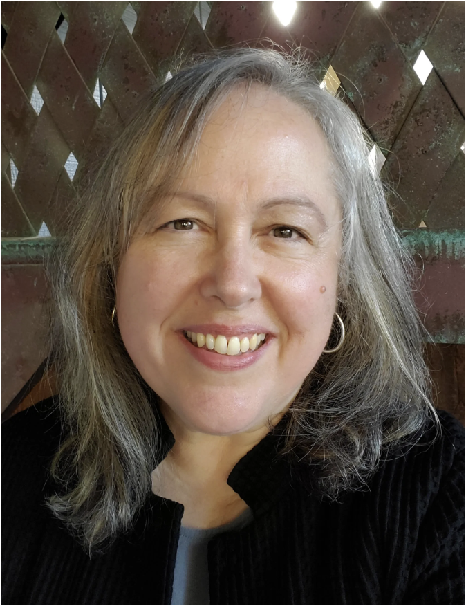 A smiling woman with shoulder-length gray hair, wearing a black top, earrings, and standing in front of a wooden lattice background.