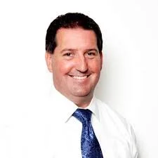 A man with dark hair, wearing a white shirt and a blue tie, smiling against a plain white background.