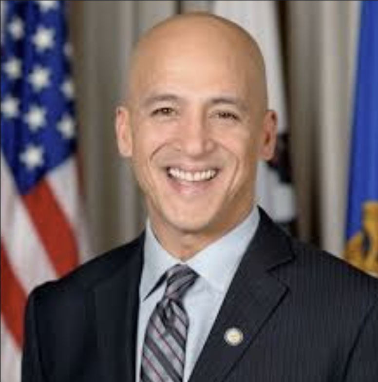 A smiling man in a suit and tie, standing in front of American flags.