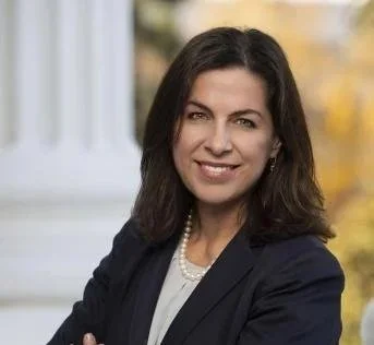 A woman with brown hair smiling and wearing a navy blazer and pearl necklace, standing outdoors with autumn trees in the background.